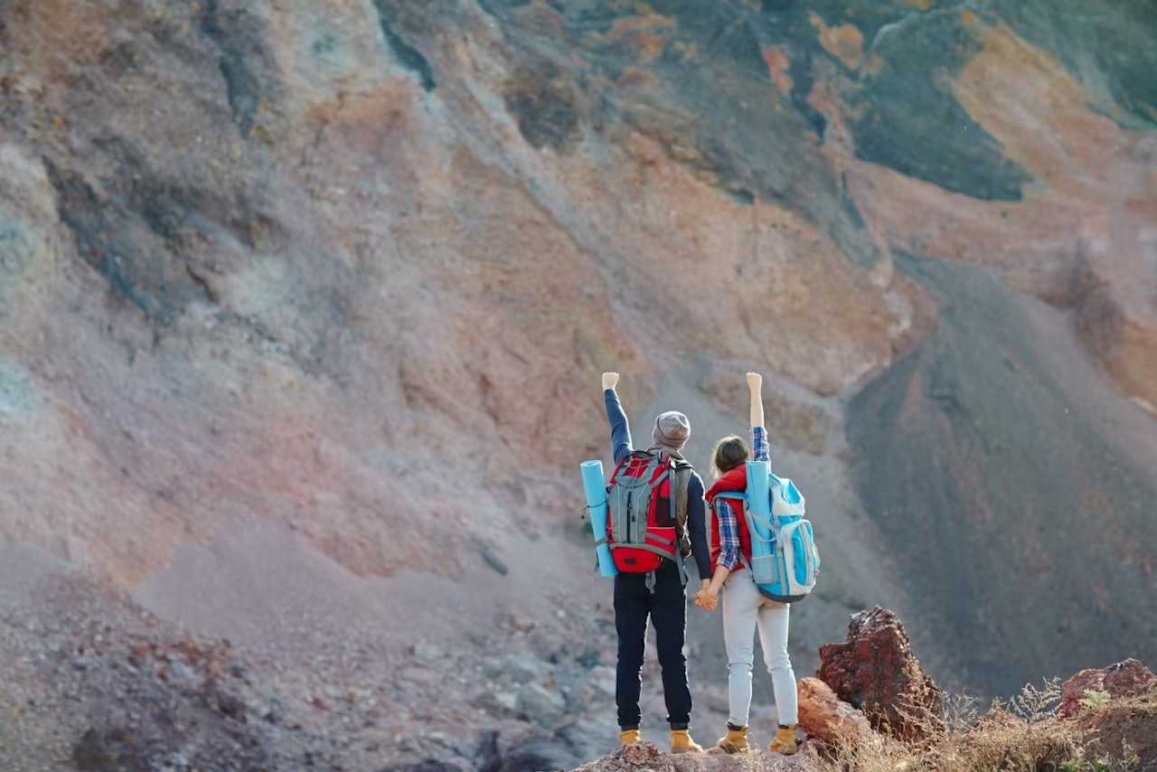 Two hikers with backpacks stand on a mountain peak, overlooking a vast landscape under a clear blue sky.