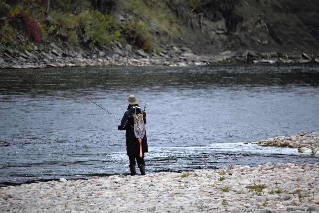 A man stands on the riverbank, casting his line into the water while fishing.