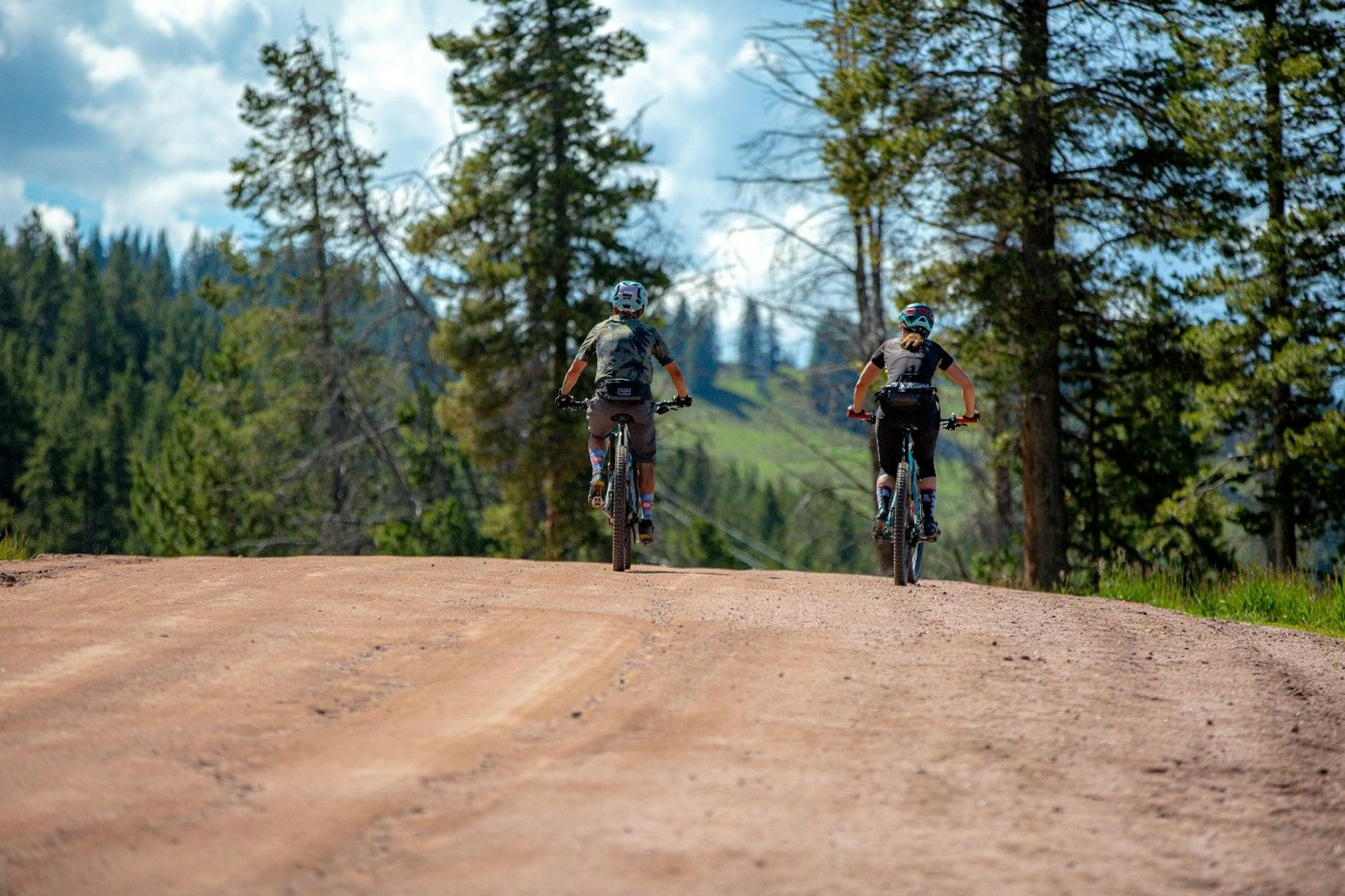 Two individuals cycling on a dirt road surrounded by greenery under a clear blue sky.