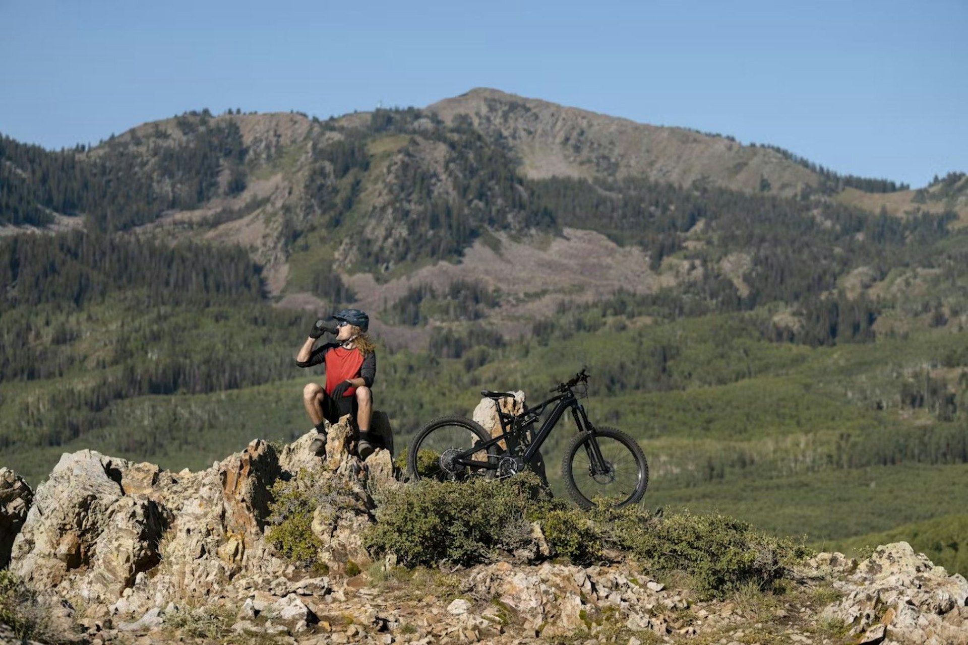 A man stands with his bike at the peak of a mountain, surrounded by a panoramic view of the landscape below.