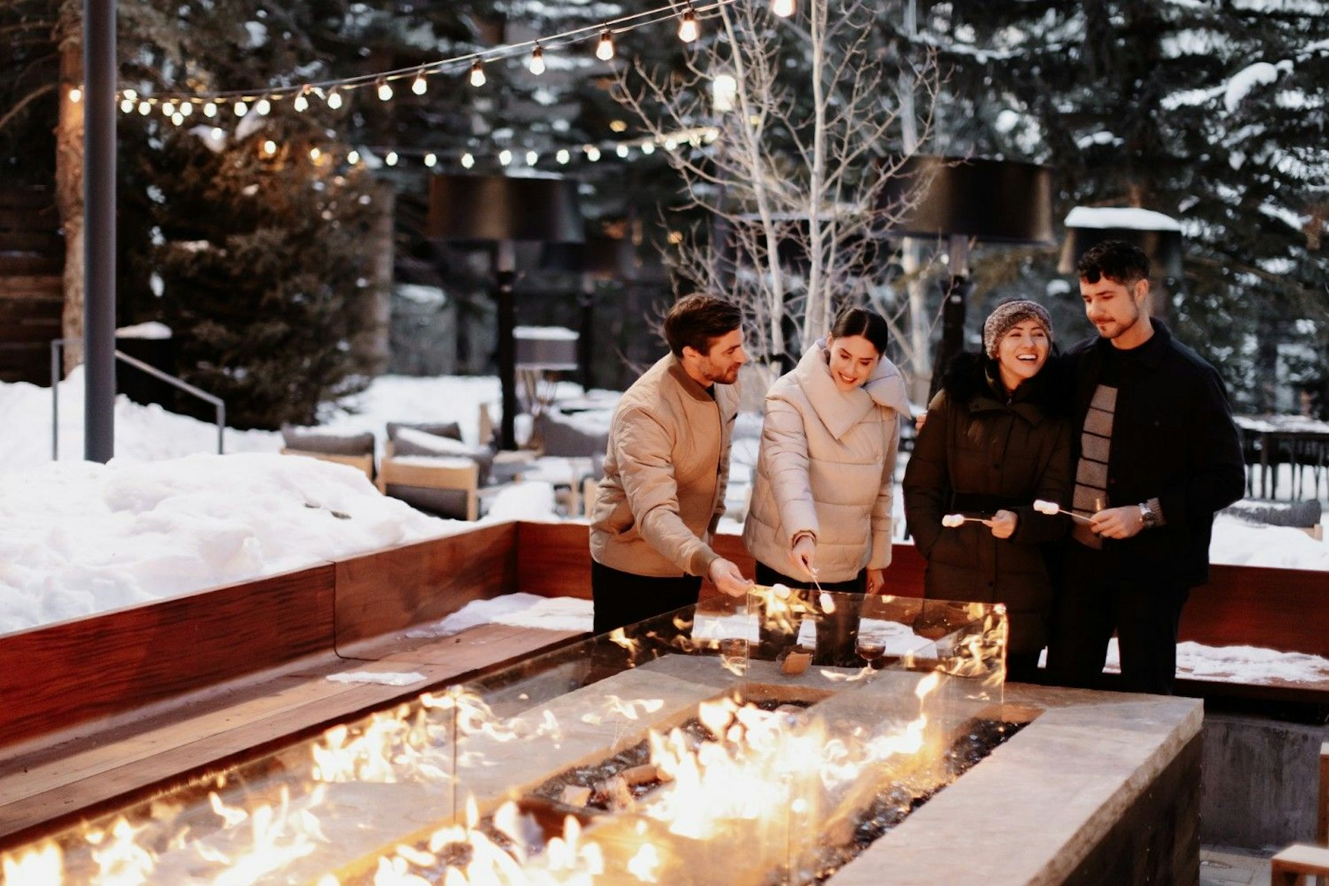 Four people gather around a fire pit, surrounded by snow, enjoying warmth and conversation in a winter setting.