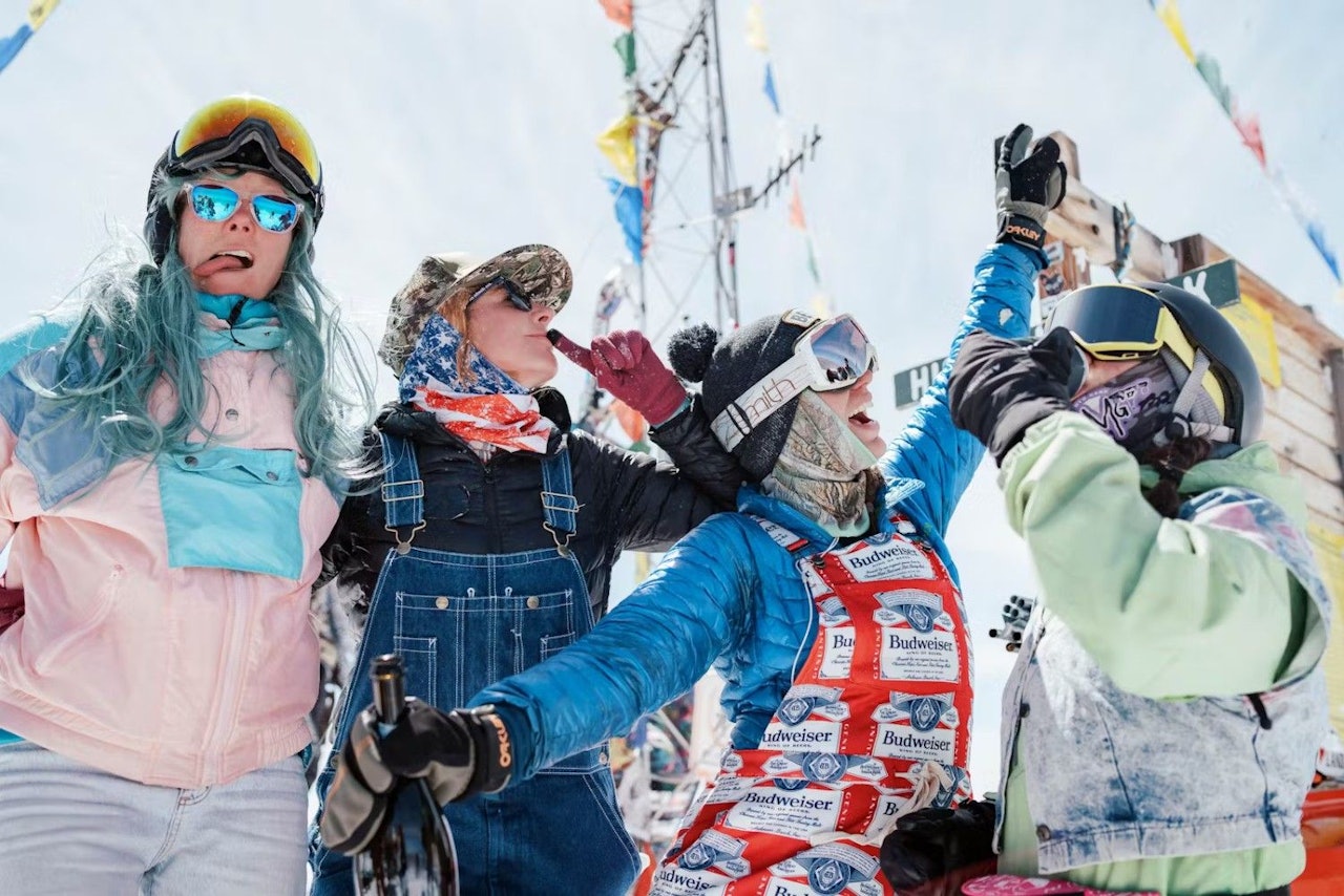 Three women in ski gear and goggles joyfully raise their hands against a snowy mountain backdrop.