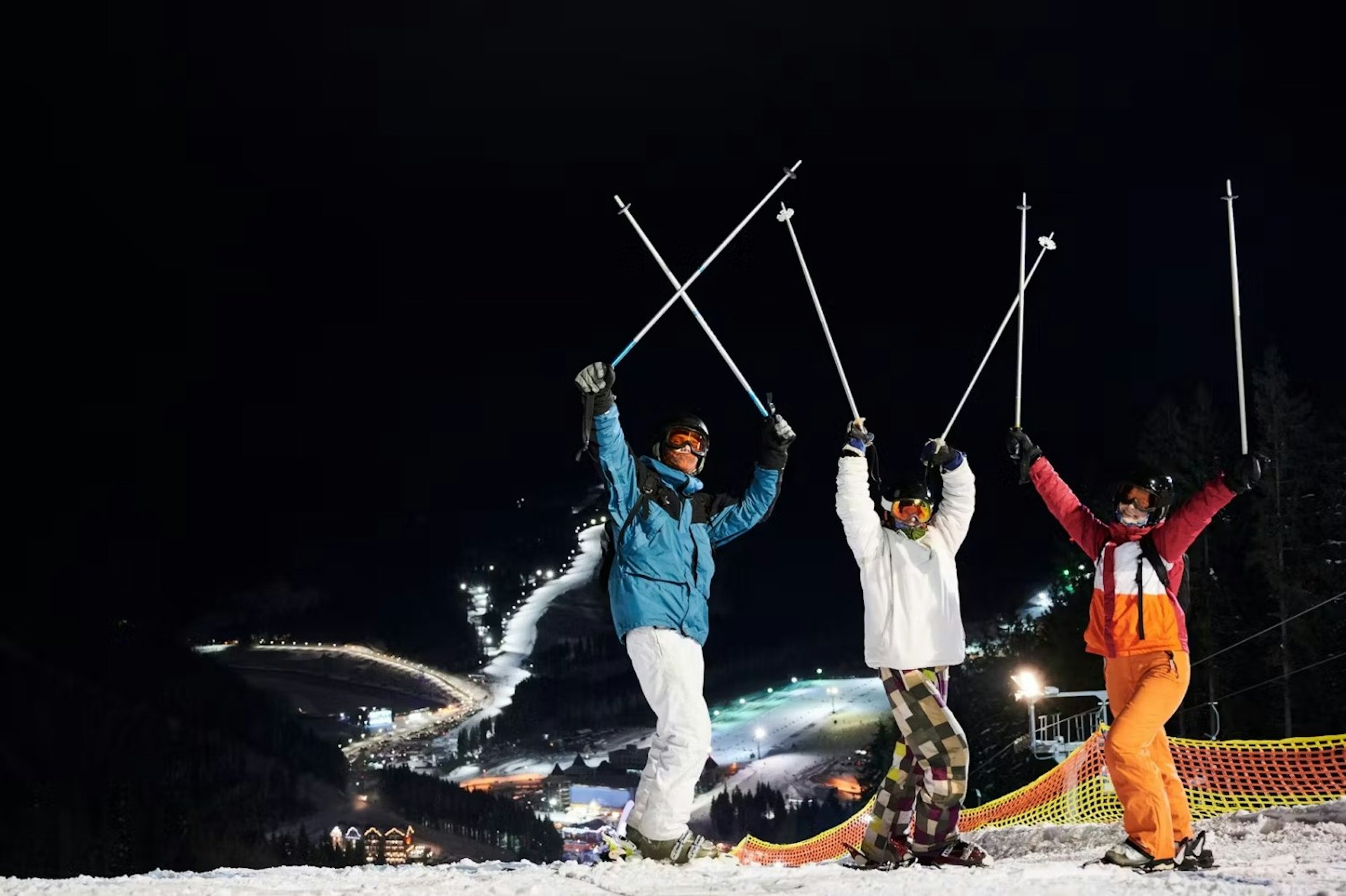 Three people raise ski poles in the air against a night sky, celebrating their skiing adventure.