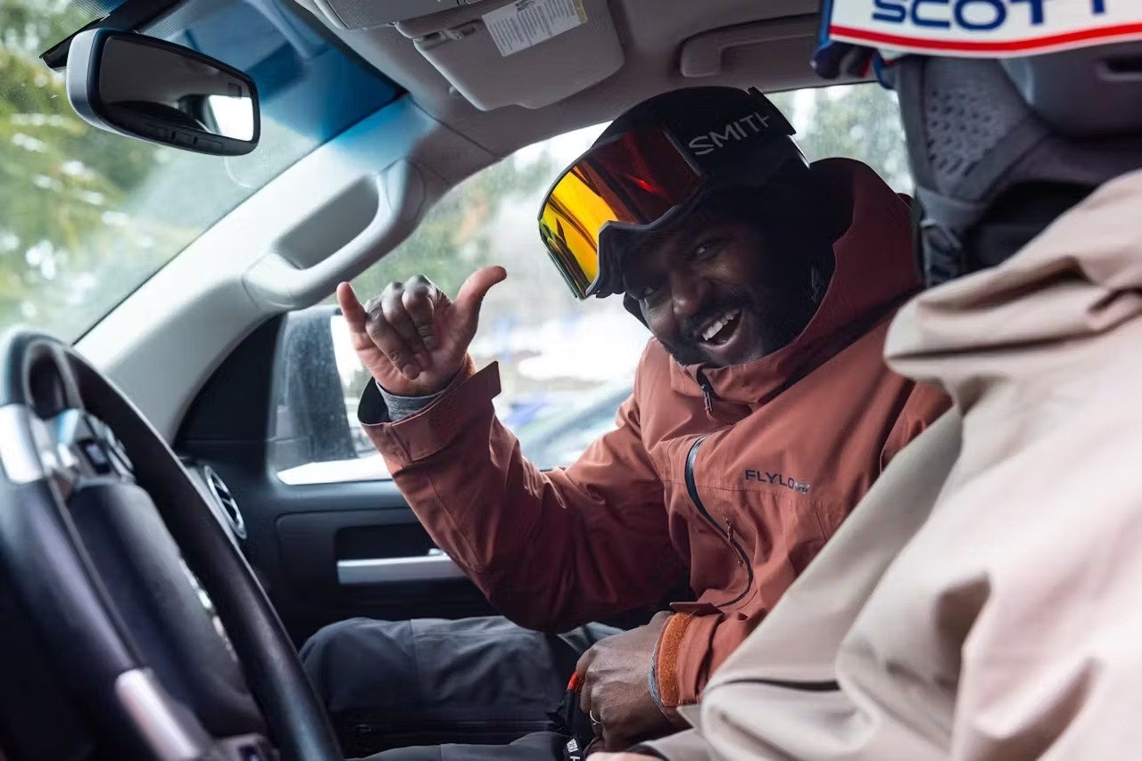 Two men in ski gear seated in the driver's seat of a car, preparing for a winter trip.