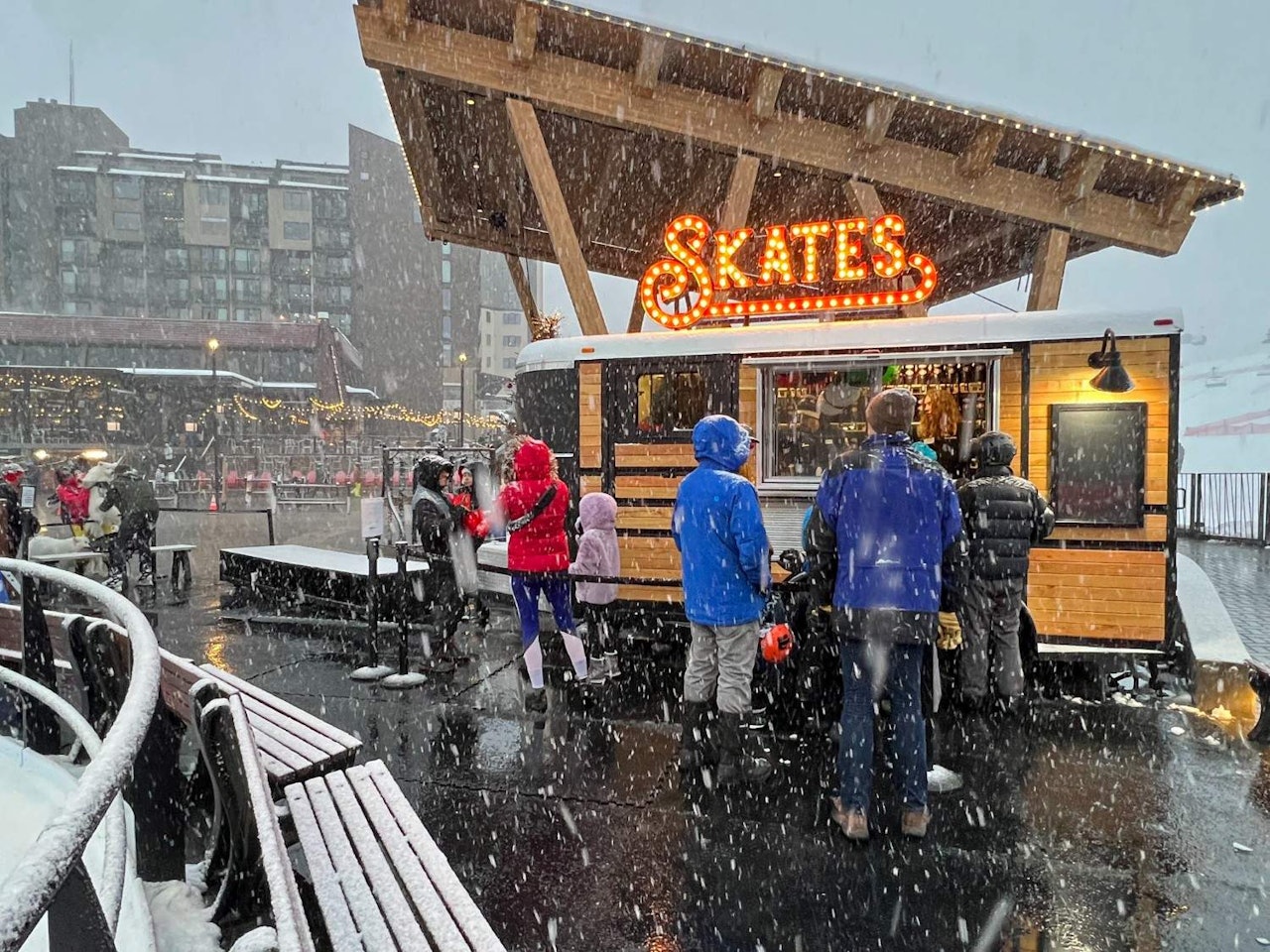 People stand outside a food truck in a snowy setting, enjoying food and drinks amidst the winter weather.