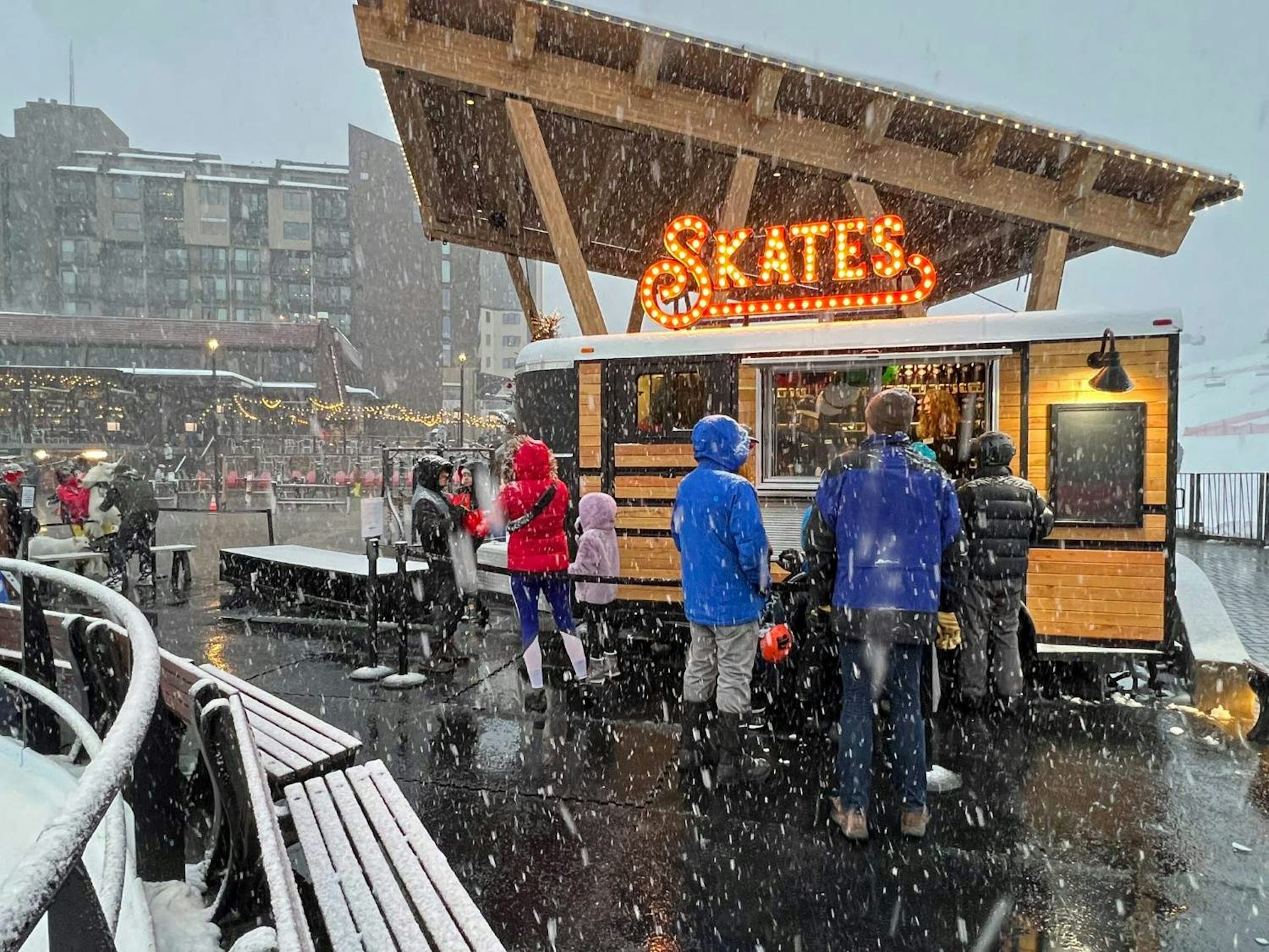 People stand outside a food truck in a snowy setting, enjoying food and drinks amidst the winter weather.