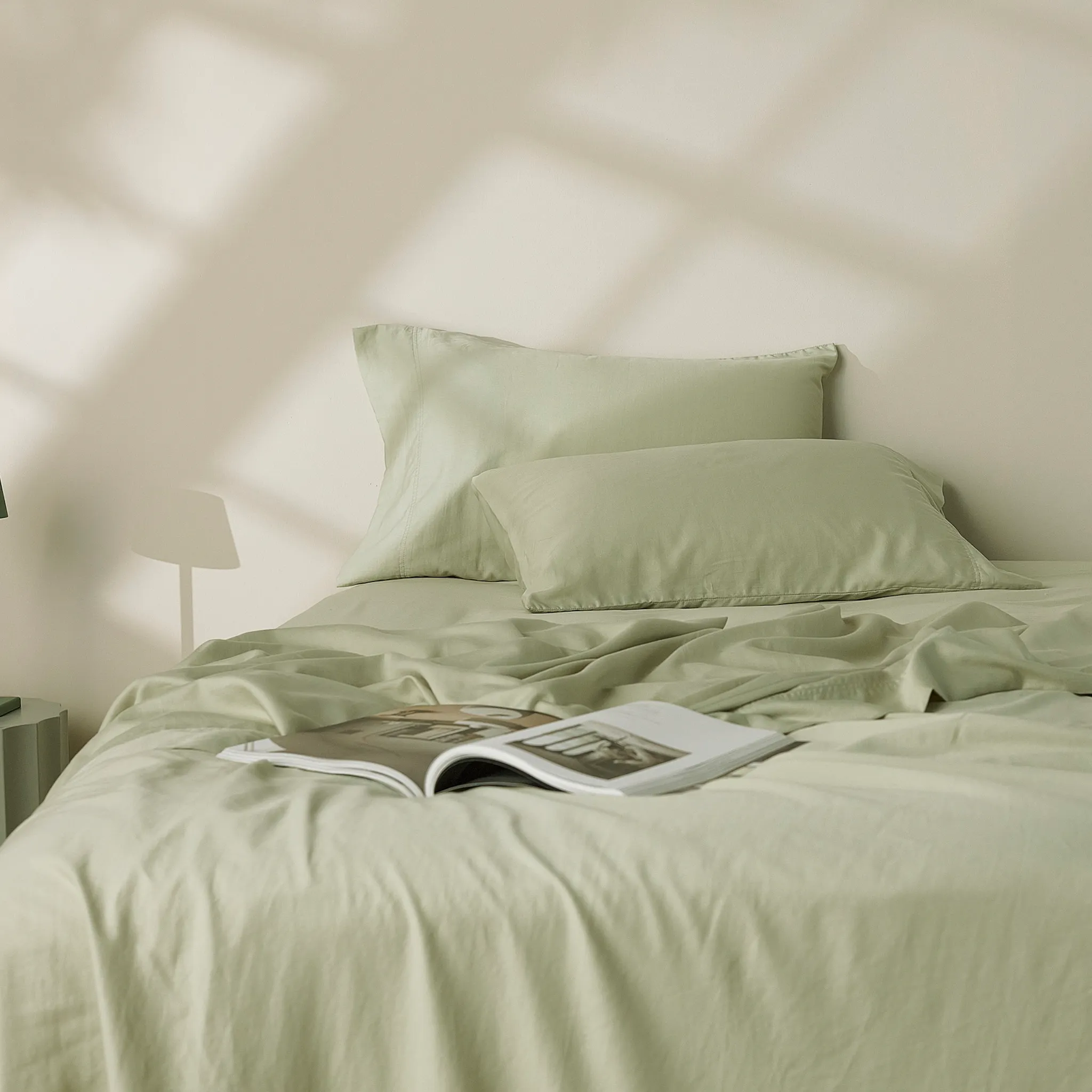 Soft sage green bedding with matching pillows and an open book on a casually layered bed in gentle sunlight.