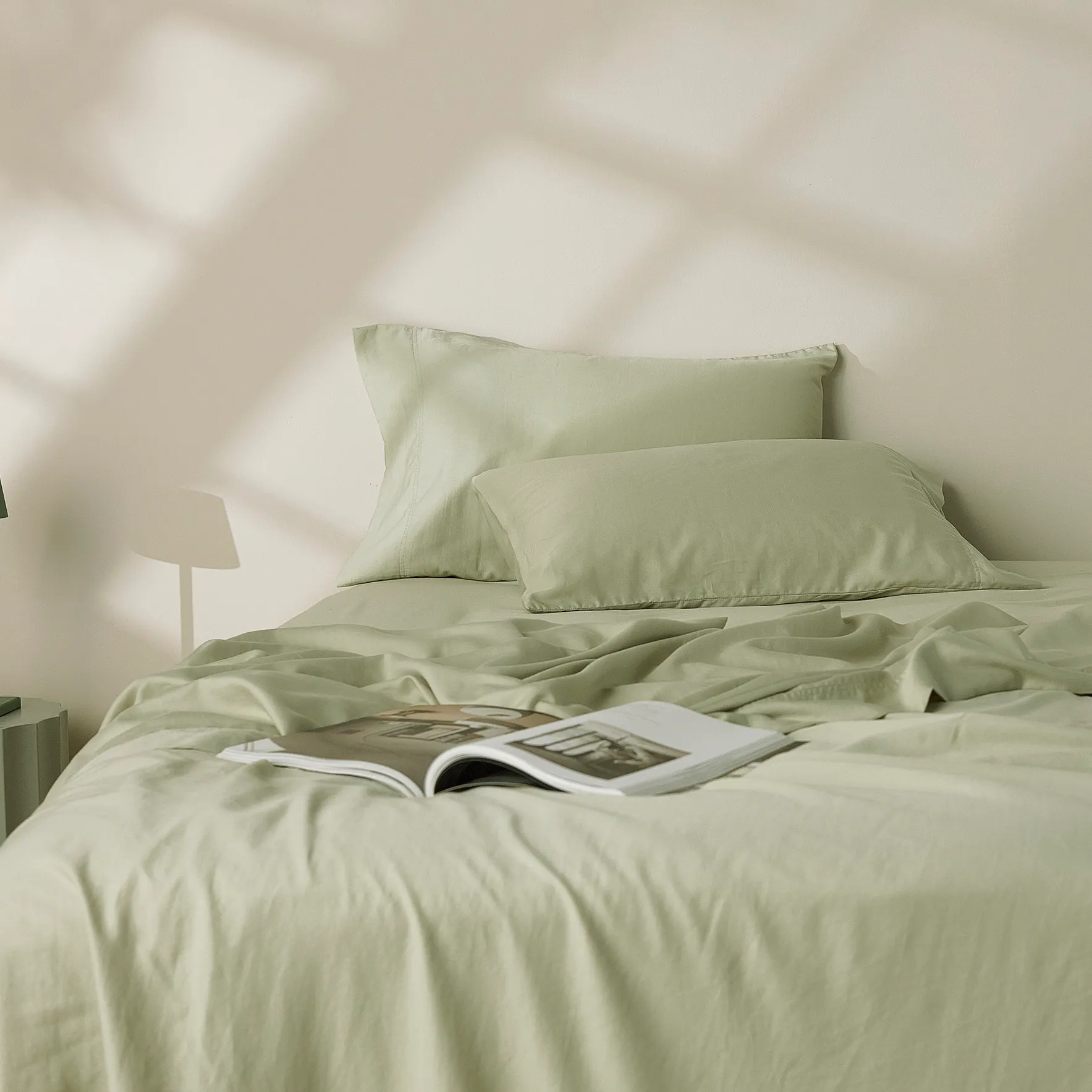 Soft sage green bedding with matching pillows and an open book on a casually layered bed in gentle sunlight.