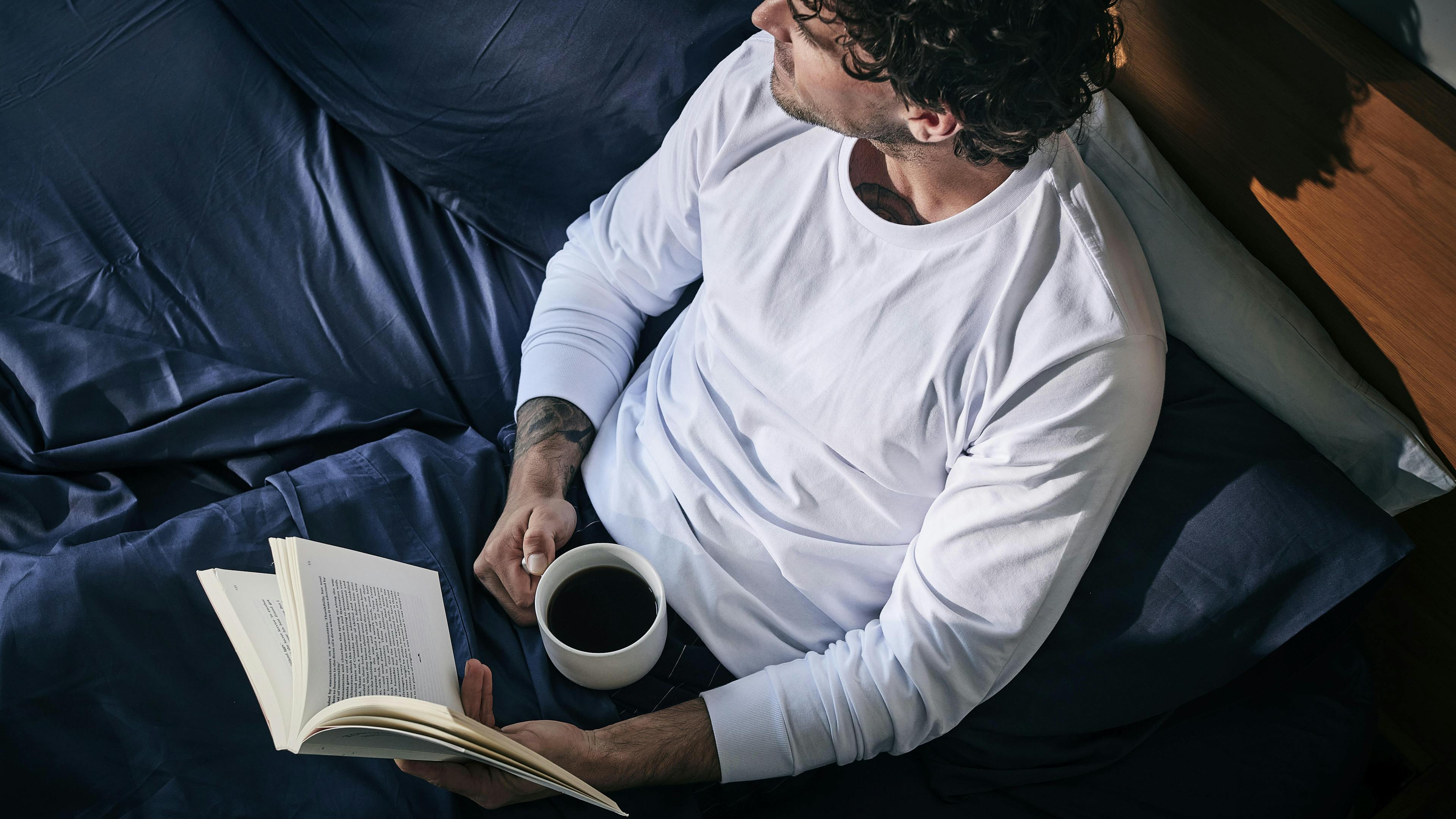 Man in blue bed sheets reading & drinking coffee