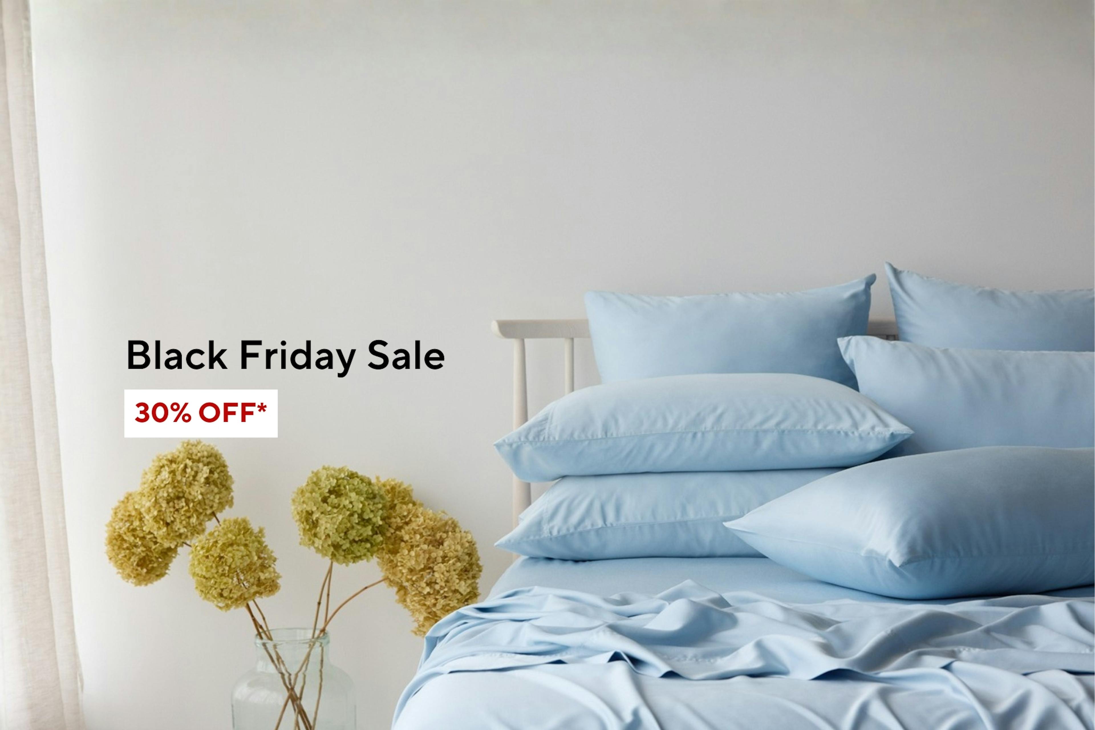 A neatly made bed with light blue sheets and pillows beside a vase of dried hydrangeas. Text on the image reads 'Black Friday Sale – 30% OFF*' in black and red letters on a white background. Shop Now Button below. 
