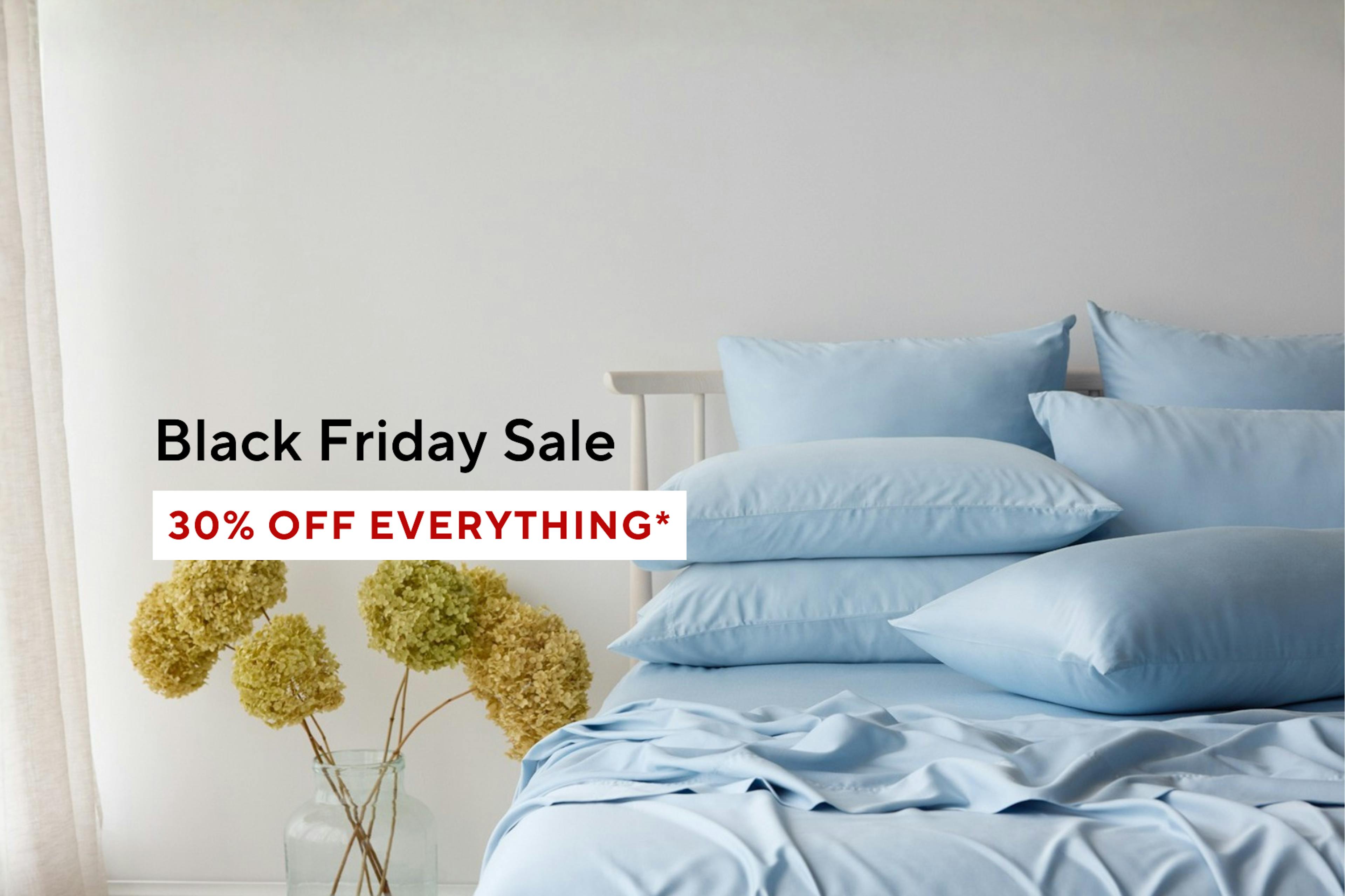 A neatly made bed with light blue sheets and pillows beside a vase of dried hydrangeas. Text on the image reads 'Black Friday Sale – 30% OFF*' in black and red letters on a white background. Shop Now Button below. 