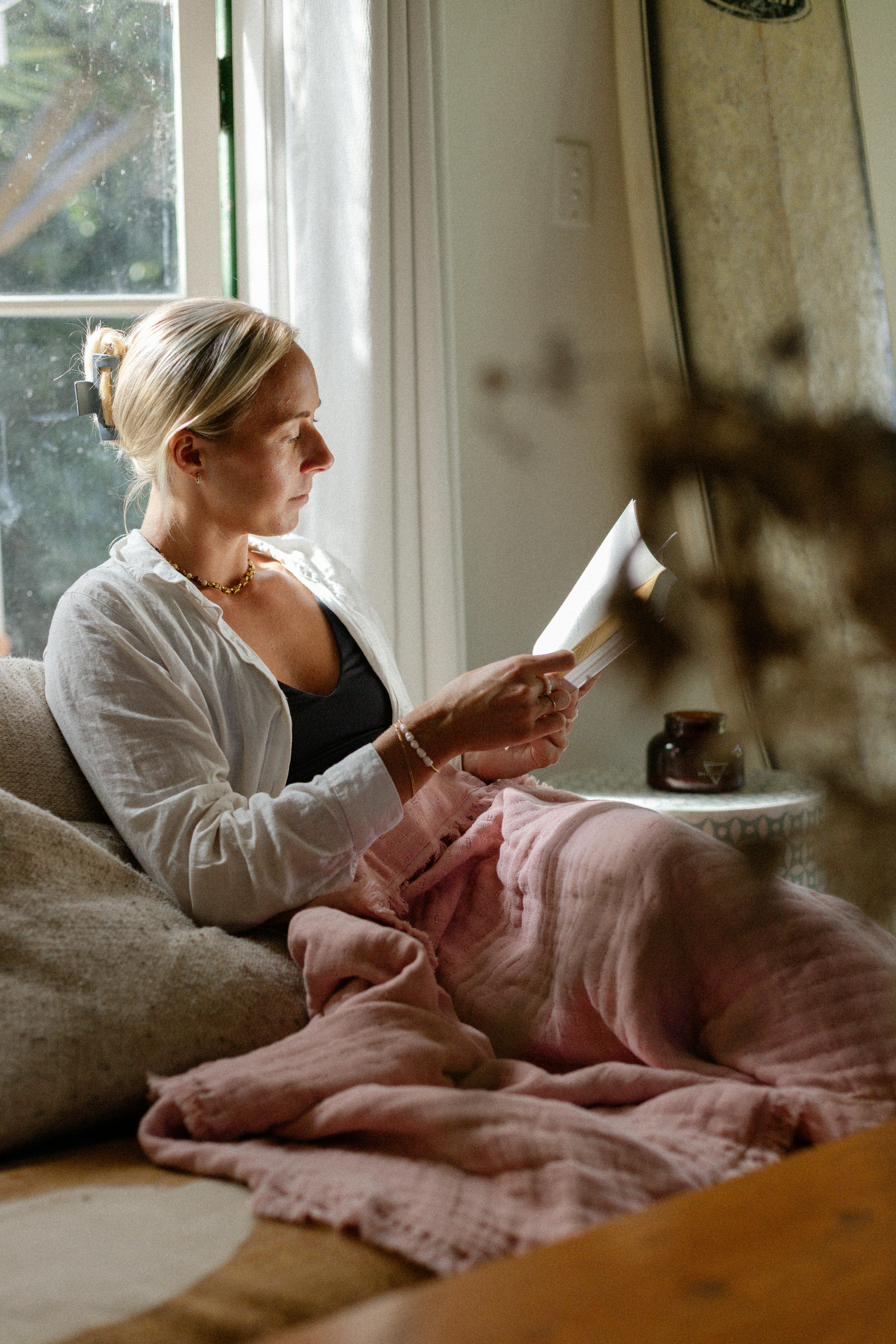 Woman relaxing on a sofa reading a book, wrapped in a soft pink blanket with natural light coming through a nearby window.