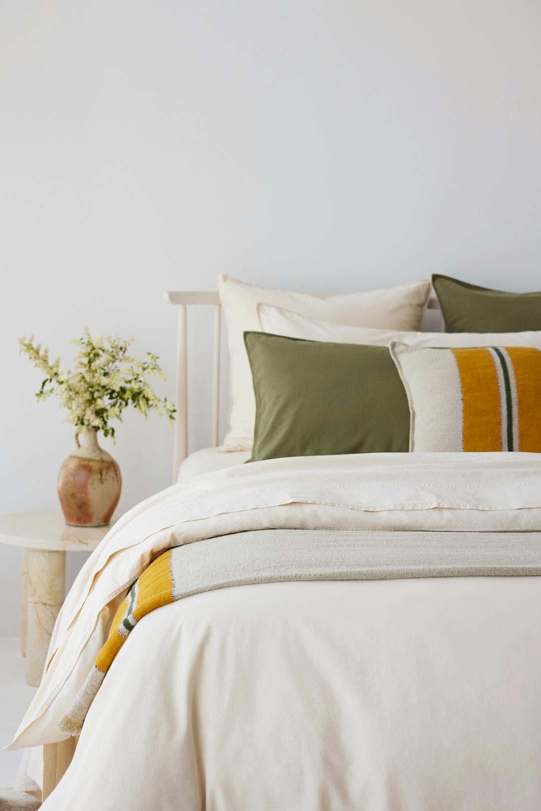 Cream bed with olive cushions and a striped throw featuring a mustard accent, next to a vase of flowers.