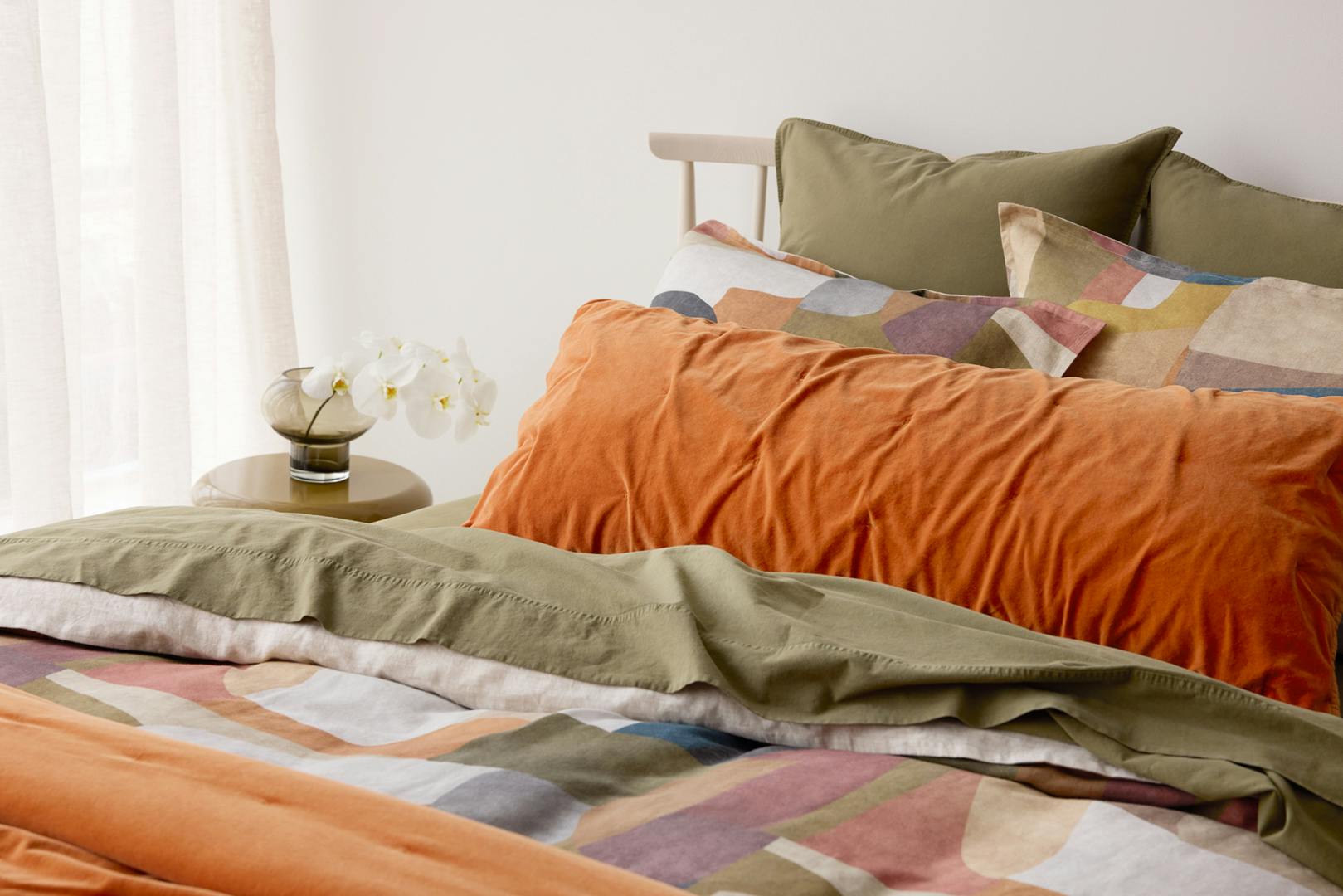Bed styled with olive and terracotta bedding and a colourful abstract duvet, with white orchids on the bedside table.
