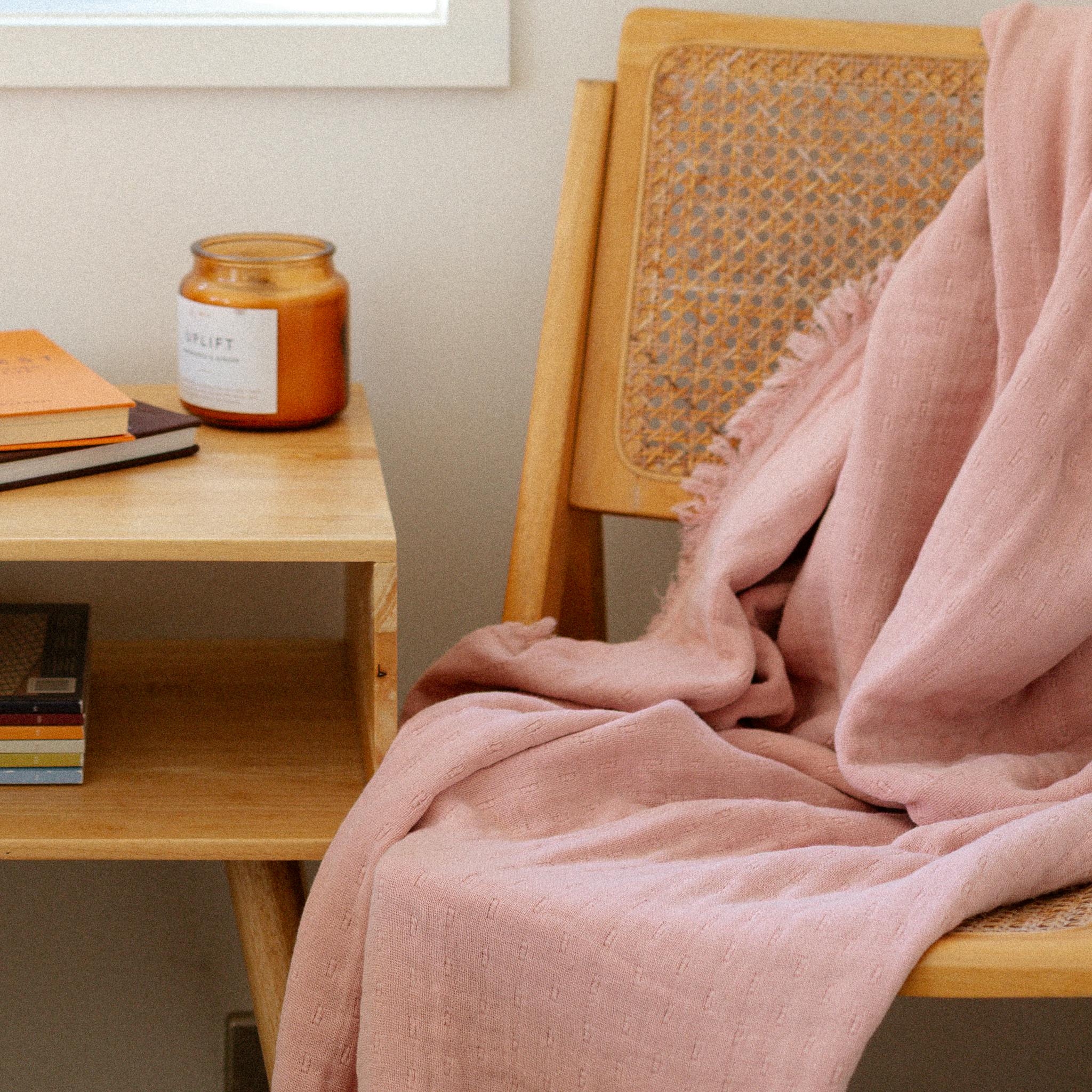 Bedroom scene with a wooden bedside table, books, a candle, and a pink throw draped over a chair by the window.