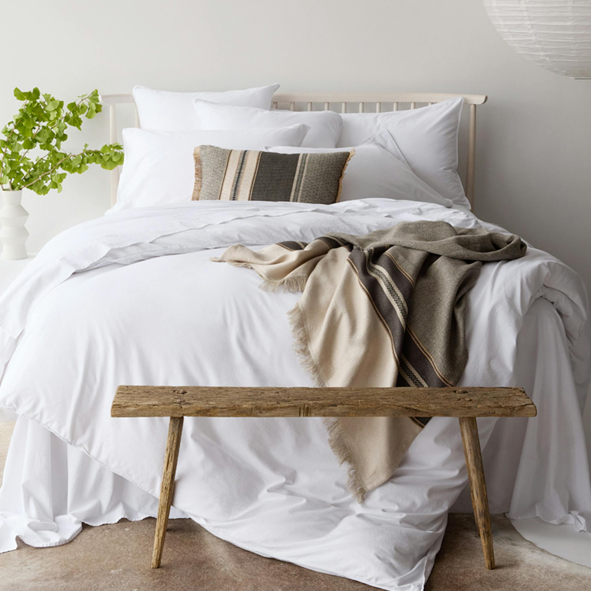 Minimal white bedroom with layered bedding, neutral throw, rustic bench, and hanging paper lantern.