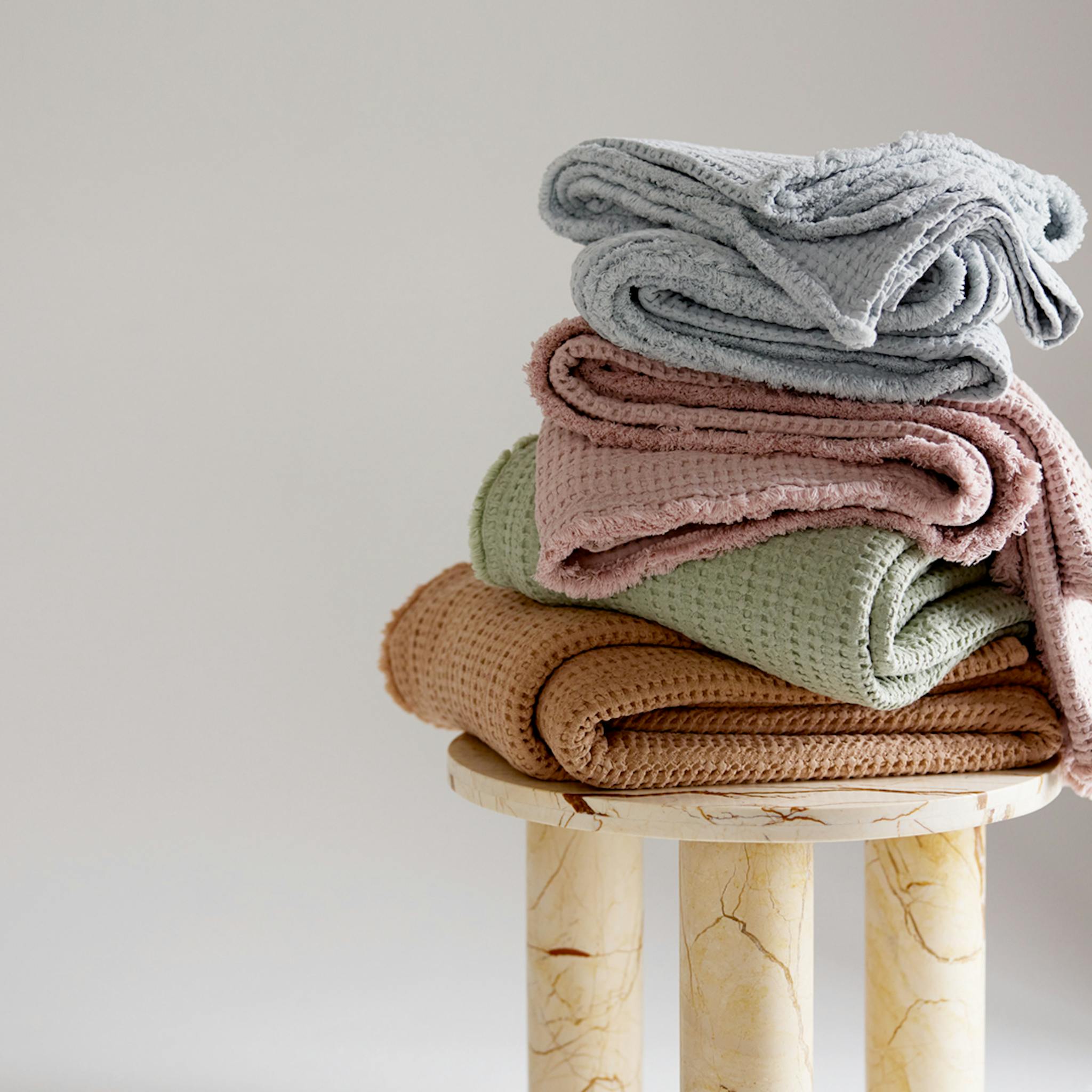 Stack of folded waffle-textured blankets in soft pastel colours on a marble stool.