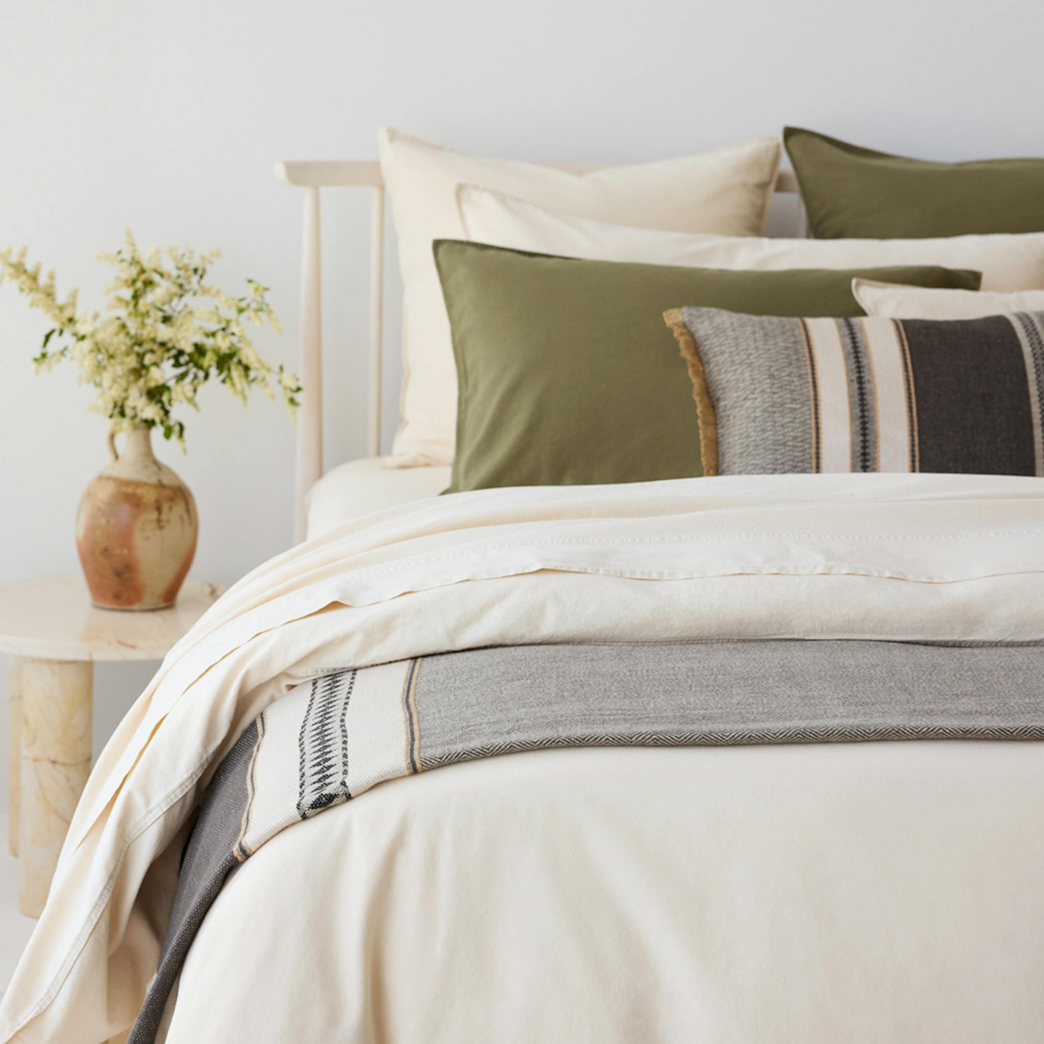 Neutral bedroom with layered cream bedding, olive cushions, and a striped throw beside a vase of flowers.