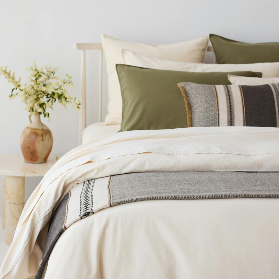 Neutral bedroom with layered cream bedding, olive cushions, and a striped throw beside a vase of flowers.