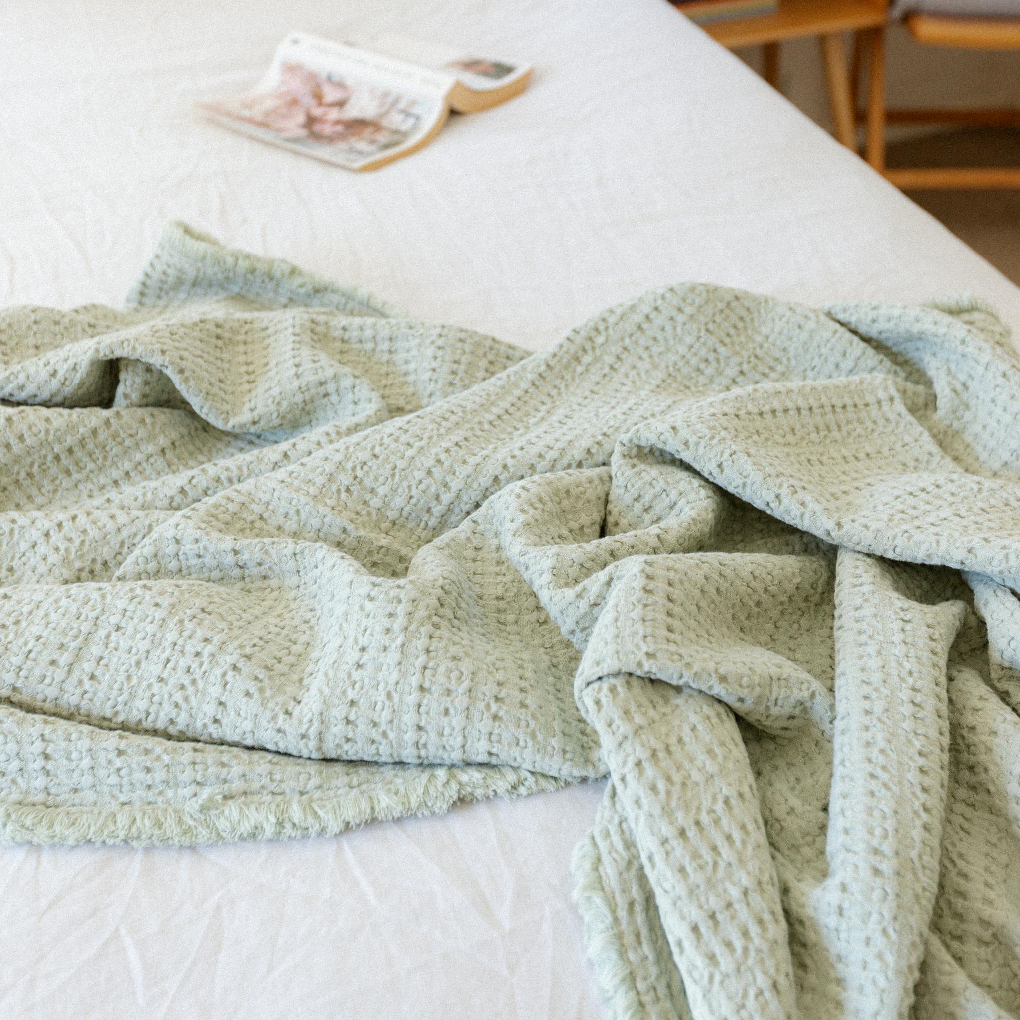 Soft green waffle blanket draped across a white bed with books and a cup on the bedside table.