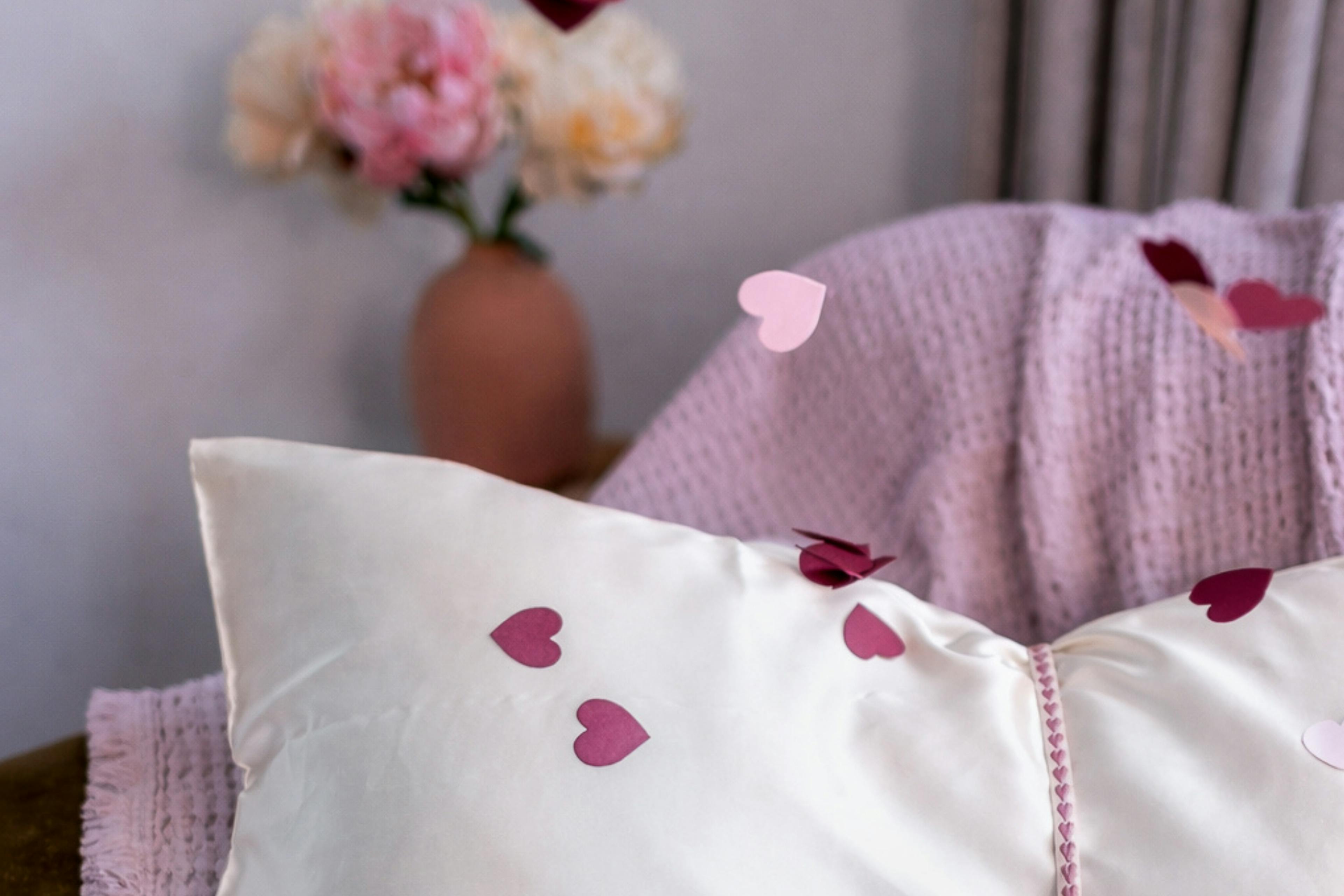 Oyster Mulberry Silk pillowcases scattered with pink and burgundy paper hearts, styled on a bed with a soft pink knitted throw and flowers in the background.