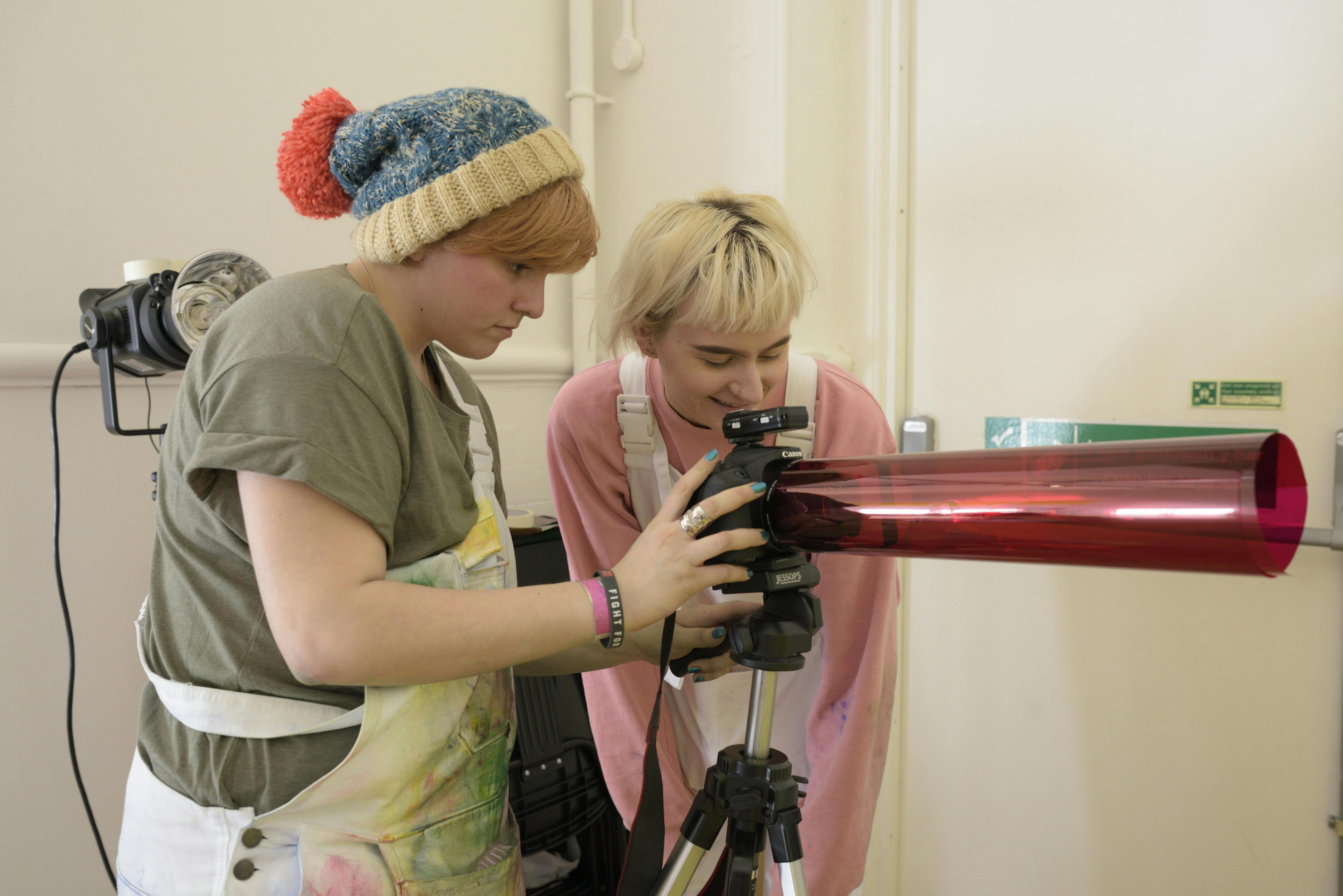 Two female students are experimenting with a camera and have wrapped a piece of semi-transparent red acetate around the lens. 