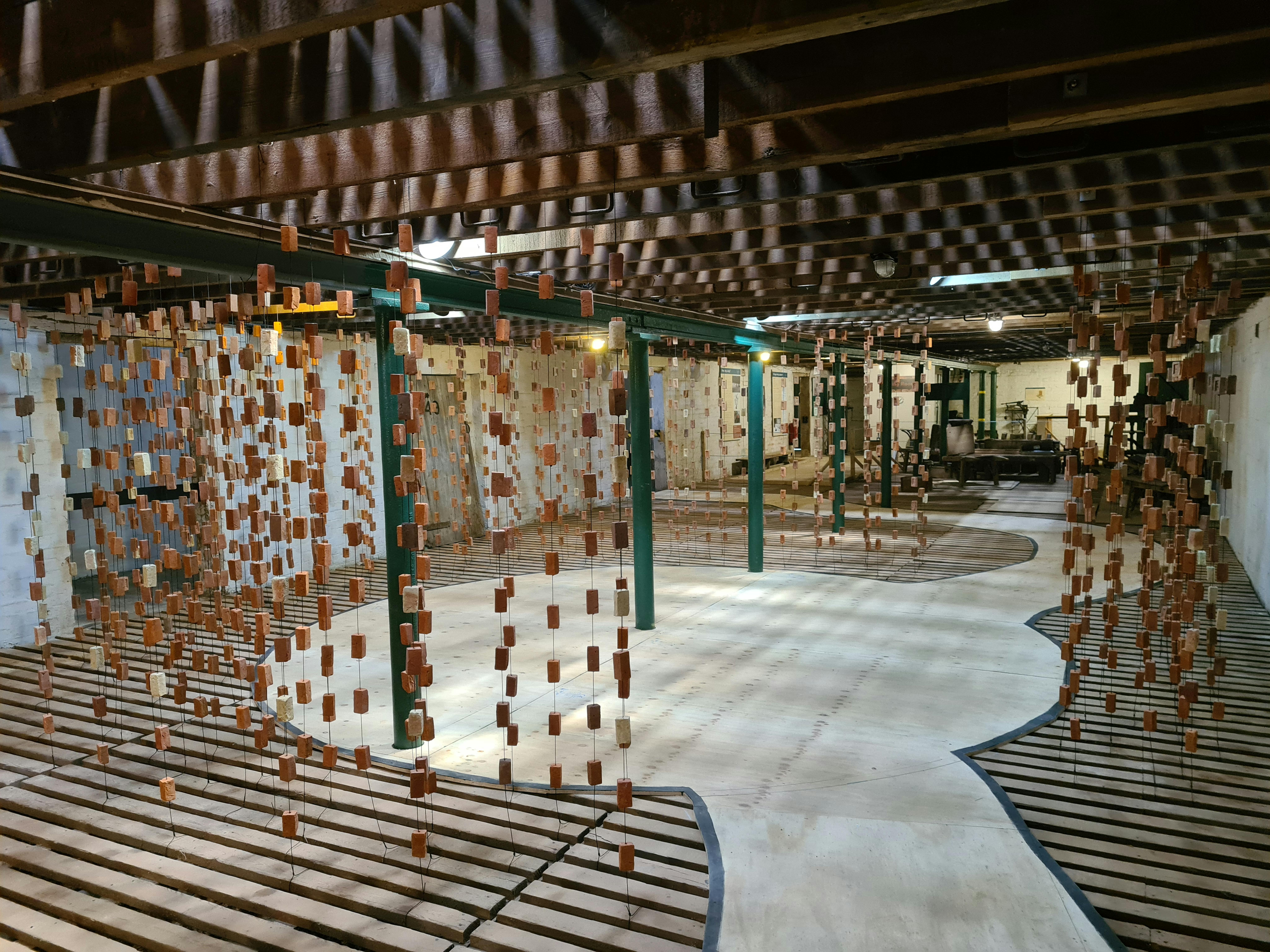 Hundreds of bricks hang from pieces of string from the ceiling. Light cascades through the room casting shadows of the bricks on the floor and walls. 