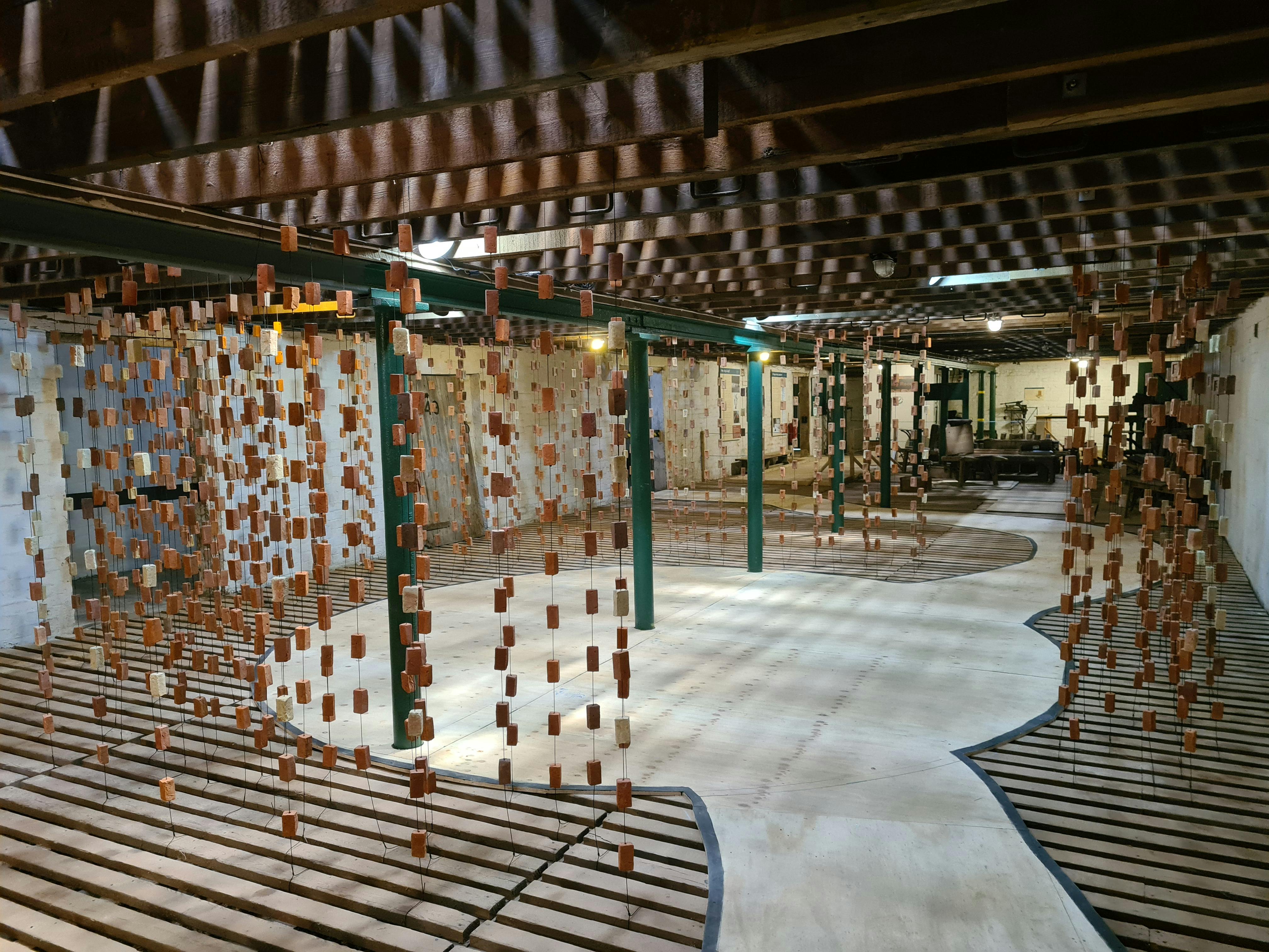 Hundreds of bricks hang from pieces of string from the ceiling. Light cascades through the room casting shadows of the bricks on the floor and walls.
