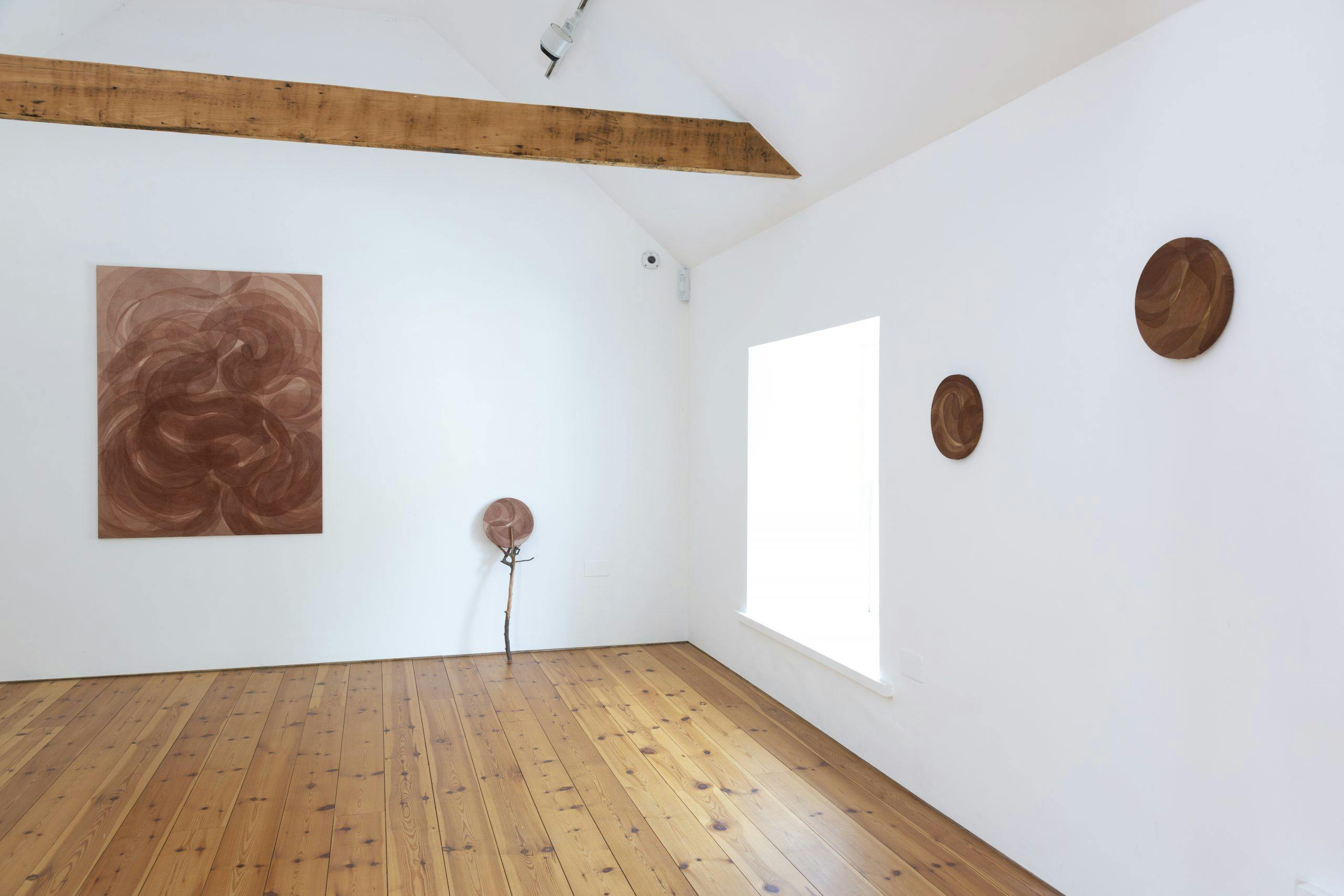 brown-scale paintings of various sizes and shapes are installed on white walls. One small round painting is propped up on a twig.