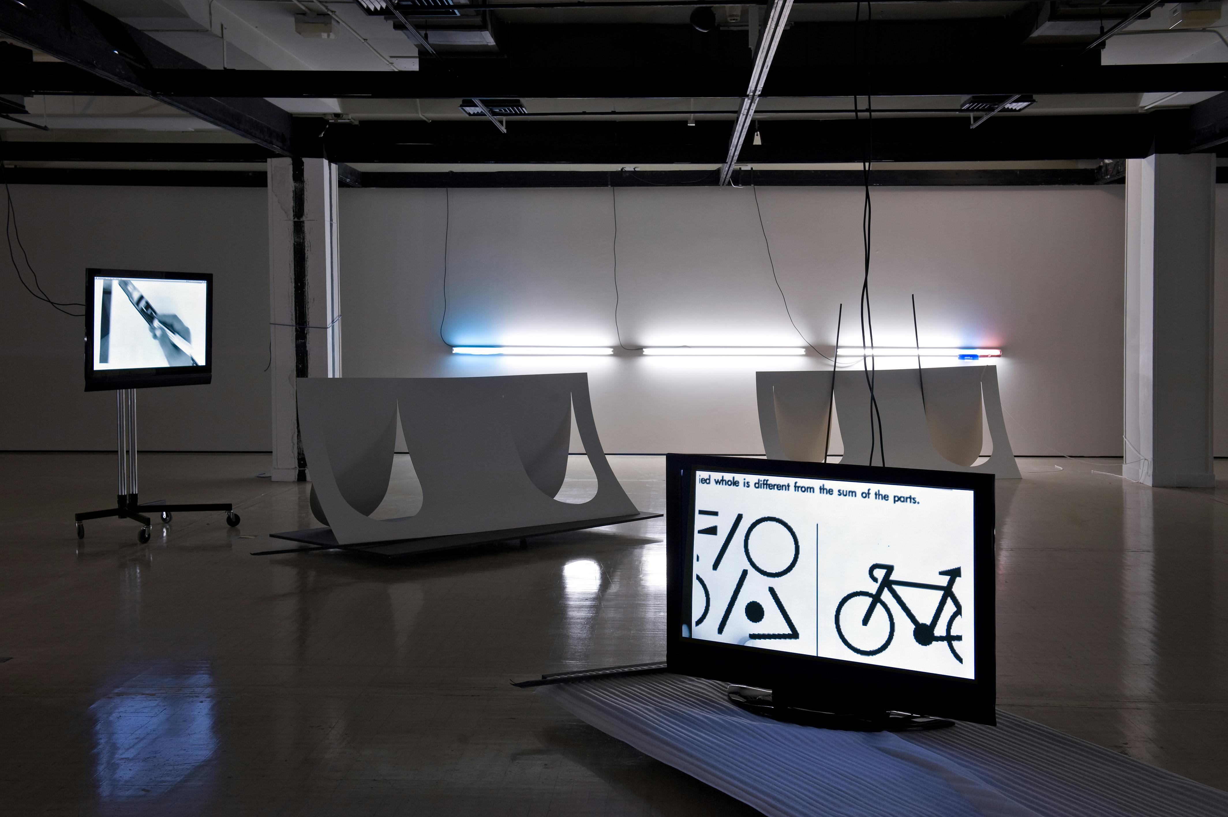 An installation in a darkened gallery. In the foreground a flat TV screen is on the floor. The background strip lighting is fixed horizontally on the wall casting strong shadows. 