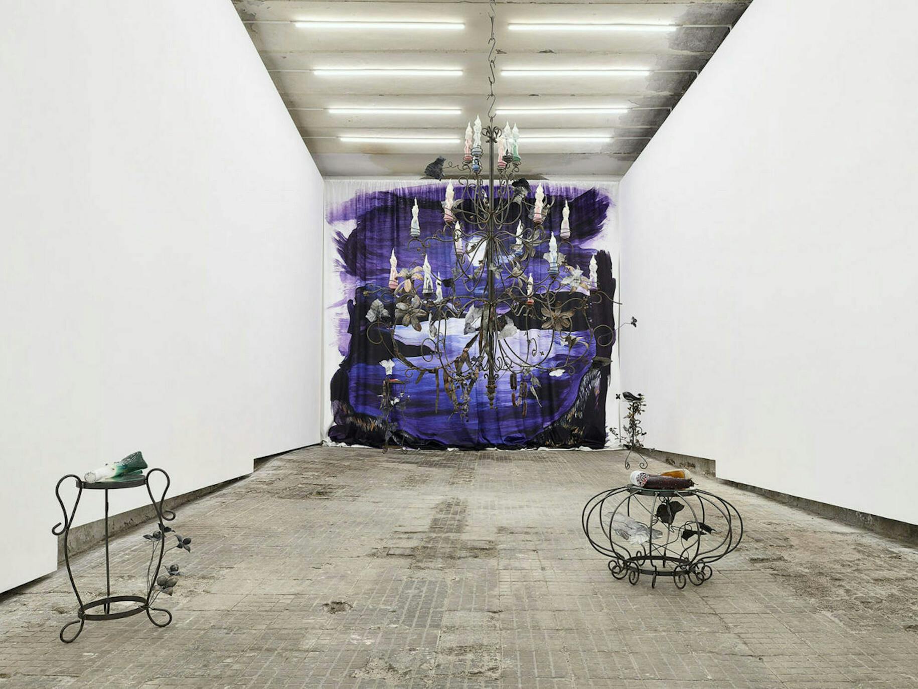 An installation in a white gallery. Small wiry sculptures made from iron sit on the floor. A large scale chandelier hangs from the ceiling with candles of naked people. A painting of a landscape is on the far wall. Everything is ornate and gothic.