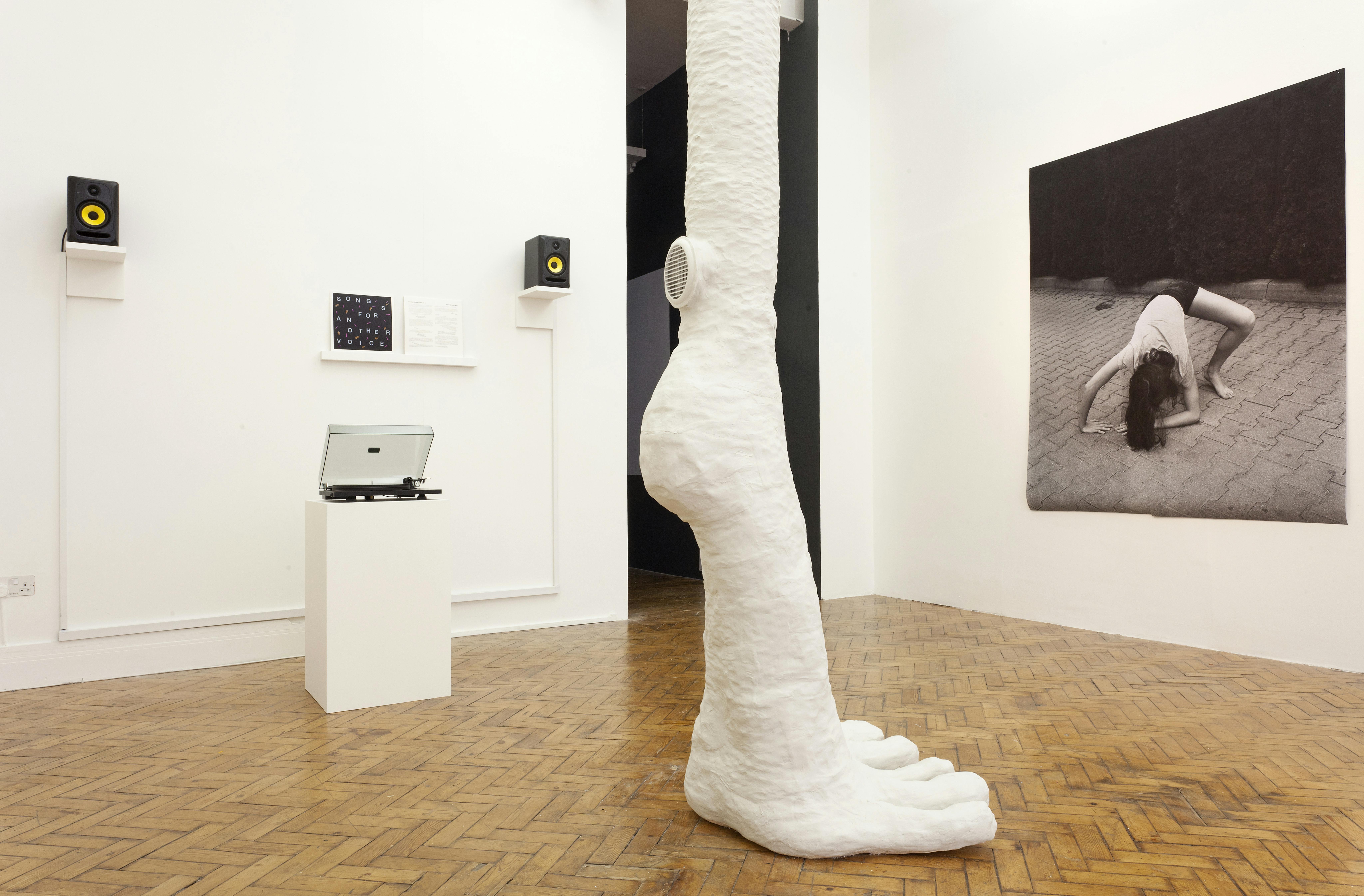 A record player is open. In the middle of the image a large foot made from plaster appears from the roof and stands on the gallery floor. On the right of the image is a large black and white photograph of a person enacting a strange uncomfortable pose. 
