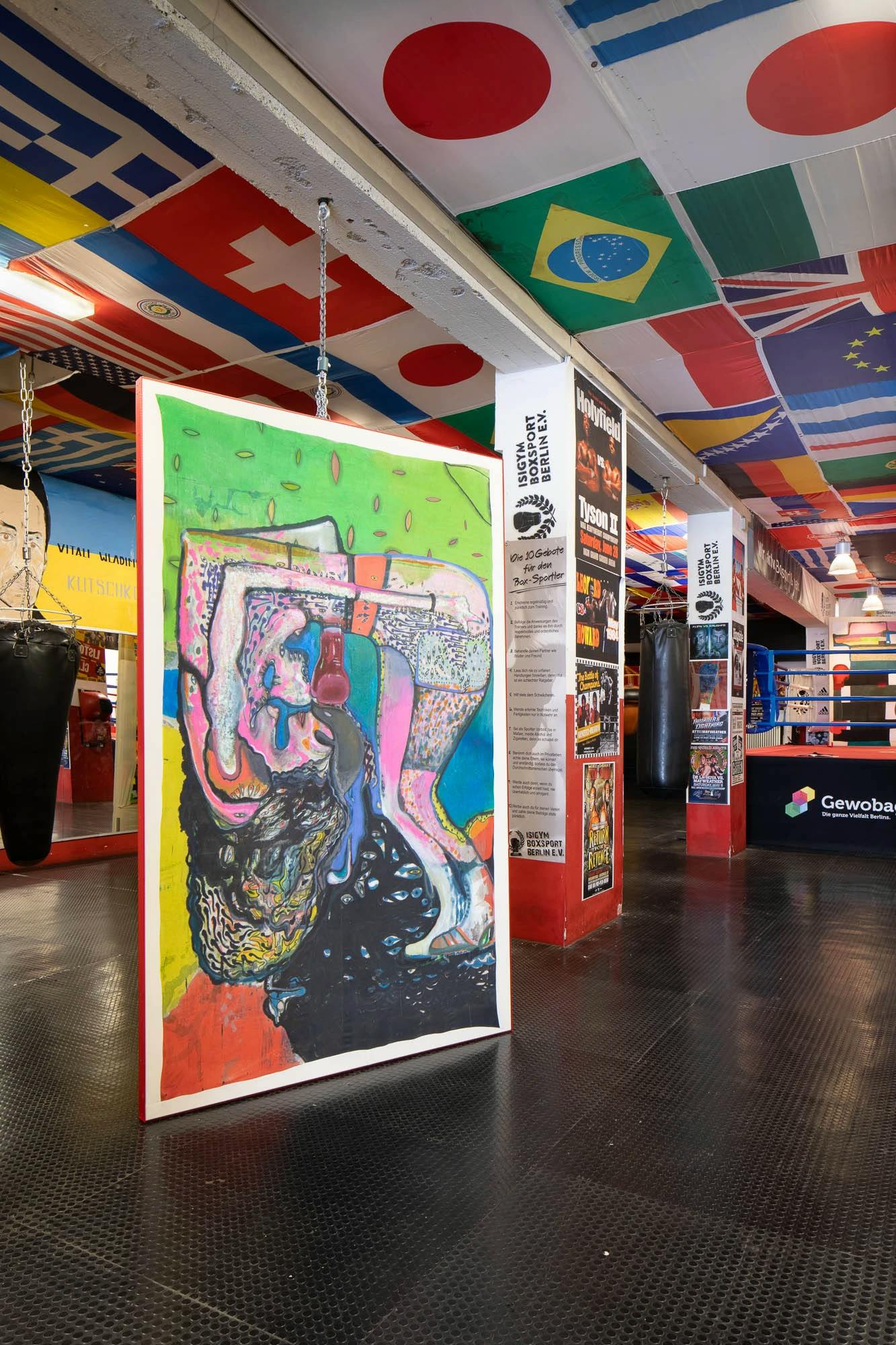 A large painting of an upside boxer in colourful tones. The painting is propped in the middle of a boxing studio surrounded by national flags and punch bags. 