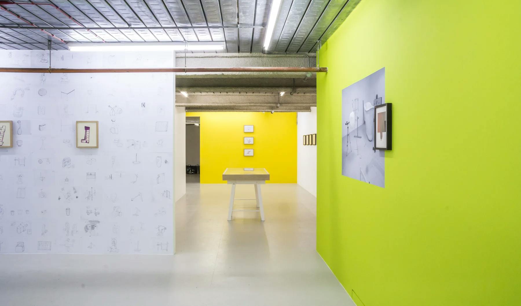 The installation frames the gallery. To the left is a wallpapered wall with drawings. In the middle is a wall painted in mustard and to the let is a wall painted in lime green. The colours are taken from the artist's notebooks. 