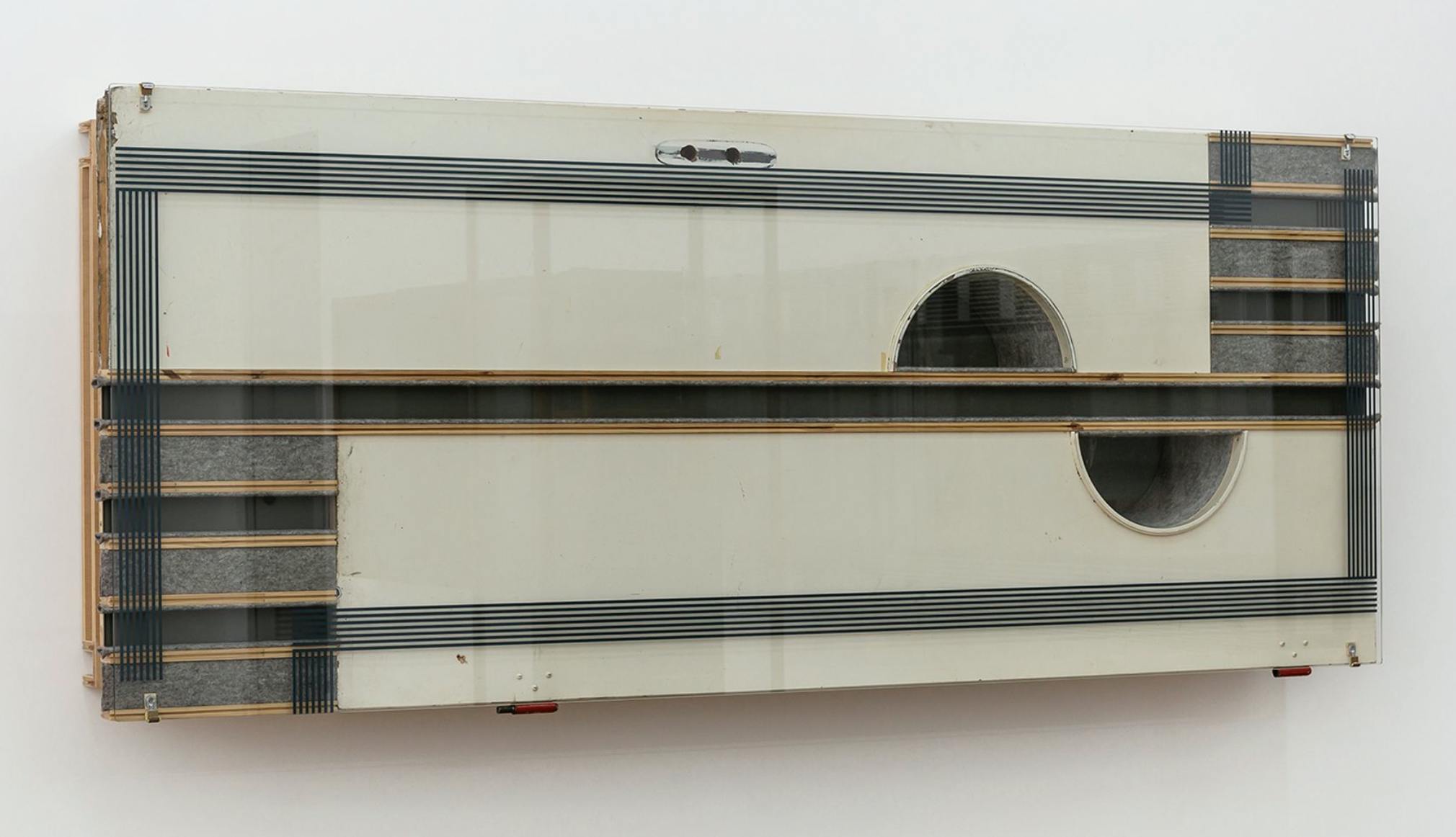 A wooden assemblage is fixed to the wall. It is a creamy colour with dark and light great accents. It has painted horizontal stripes across the surface. The wooden form is made by different parts of found wooden furniture that have been cut into abstract shapes including circles and squares.