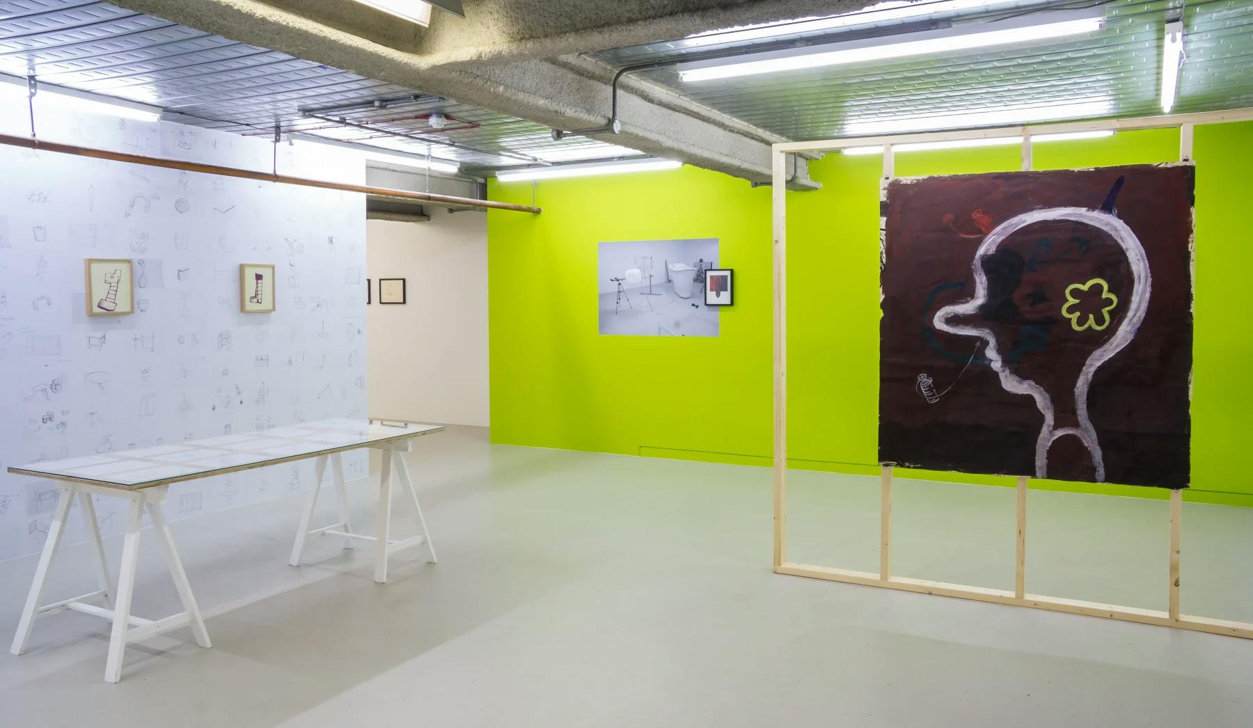 To the left is a series of artist's drawings and doodles photocopied and pasted to the wall. To the right is a large painting of a cartoon-like head with long nose installed on a timber frame. 