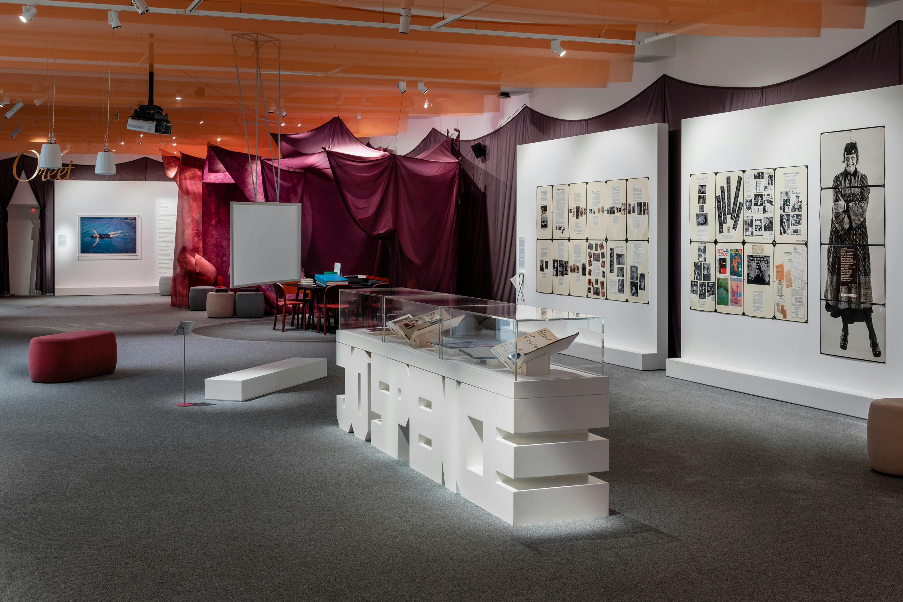 The installation includes large photographs and montages of Jo Spence on white walls. In the foreground of the image a vitrine is shaped in Jo Spence's name and contains notebooks and diaries. The gallery walls and ceiling are covered in peach coloured and dark brown fabrics creating a comforting environment.