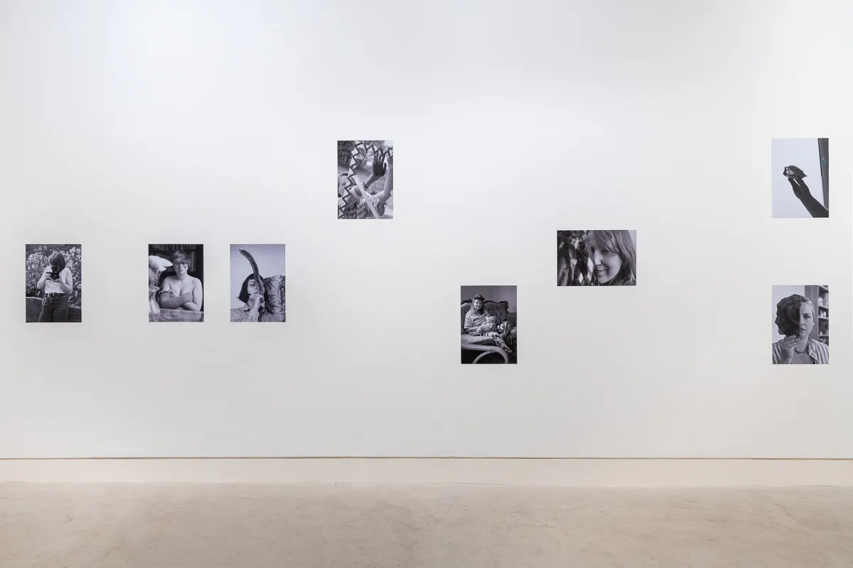 A bright white wall is covered in black and white photographs of female artists doing everyday activities.