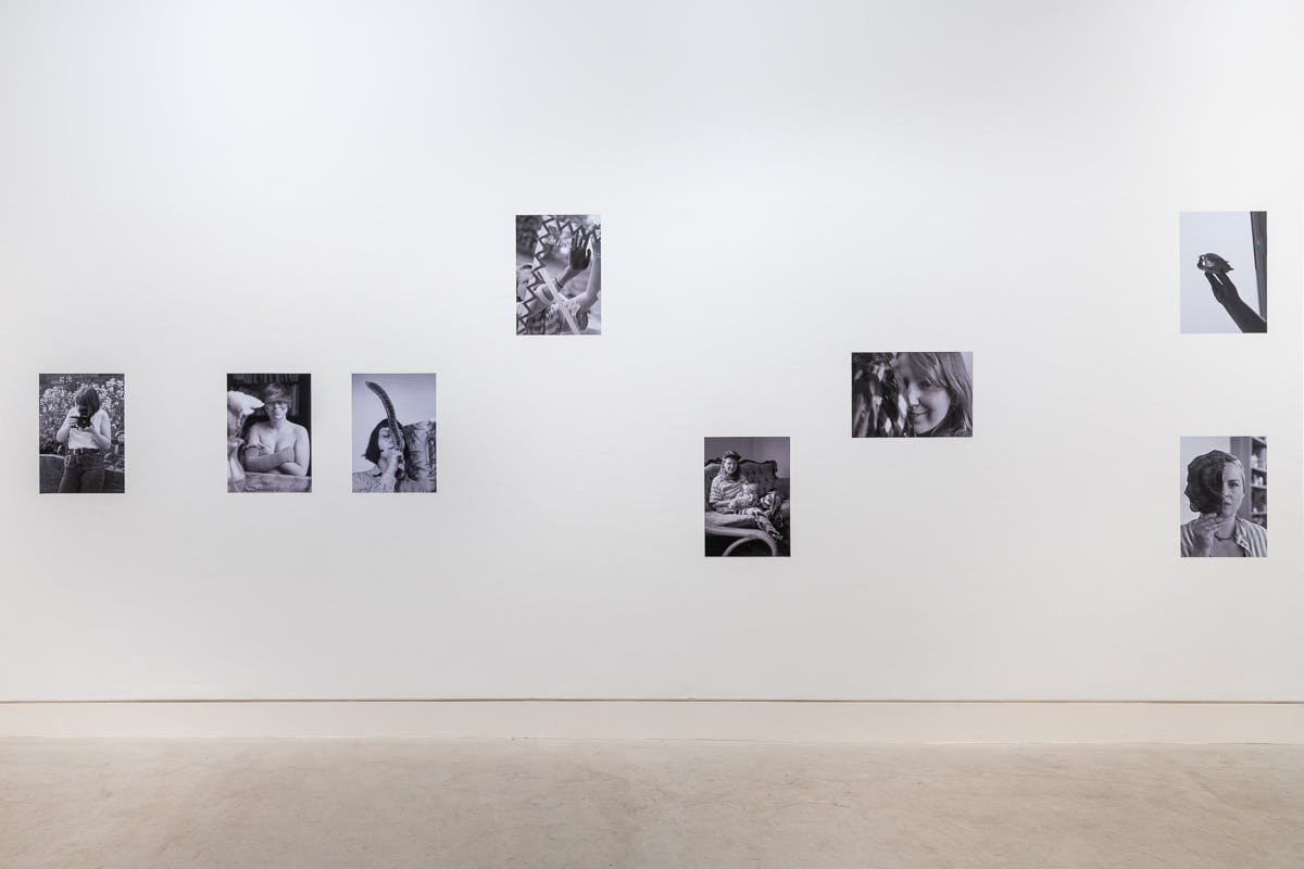 A bright white wall is covered in black and white photographs of female artists doing everyday activities. 