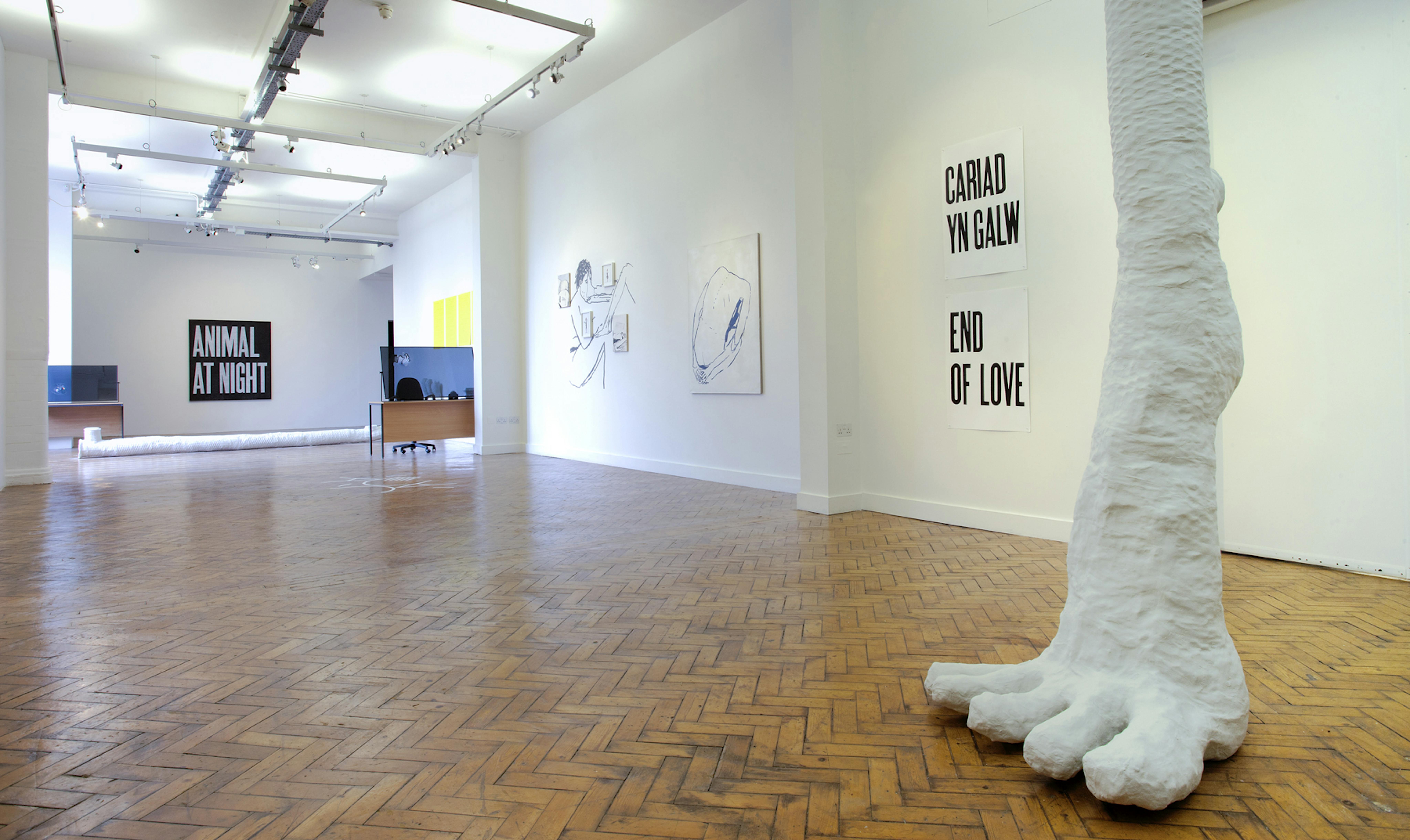 A large white foot appears from the ceiling of the gallery. It frames a long deep photograph depicting the installation of an exhibition with lots of paintings and drawings on the wall. 
