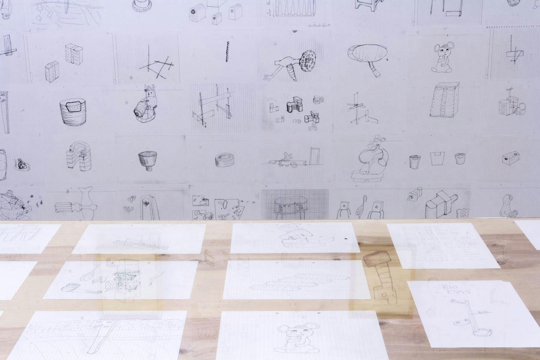 A wall with pasted drawings from the artist's studio frame a series of smaller drawings that express ideas for potential sculptures that use everyday objects like furniture and children's toys. 