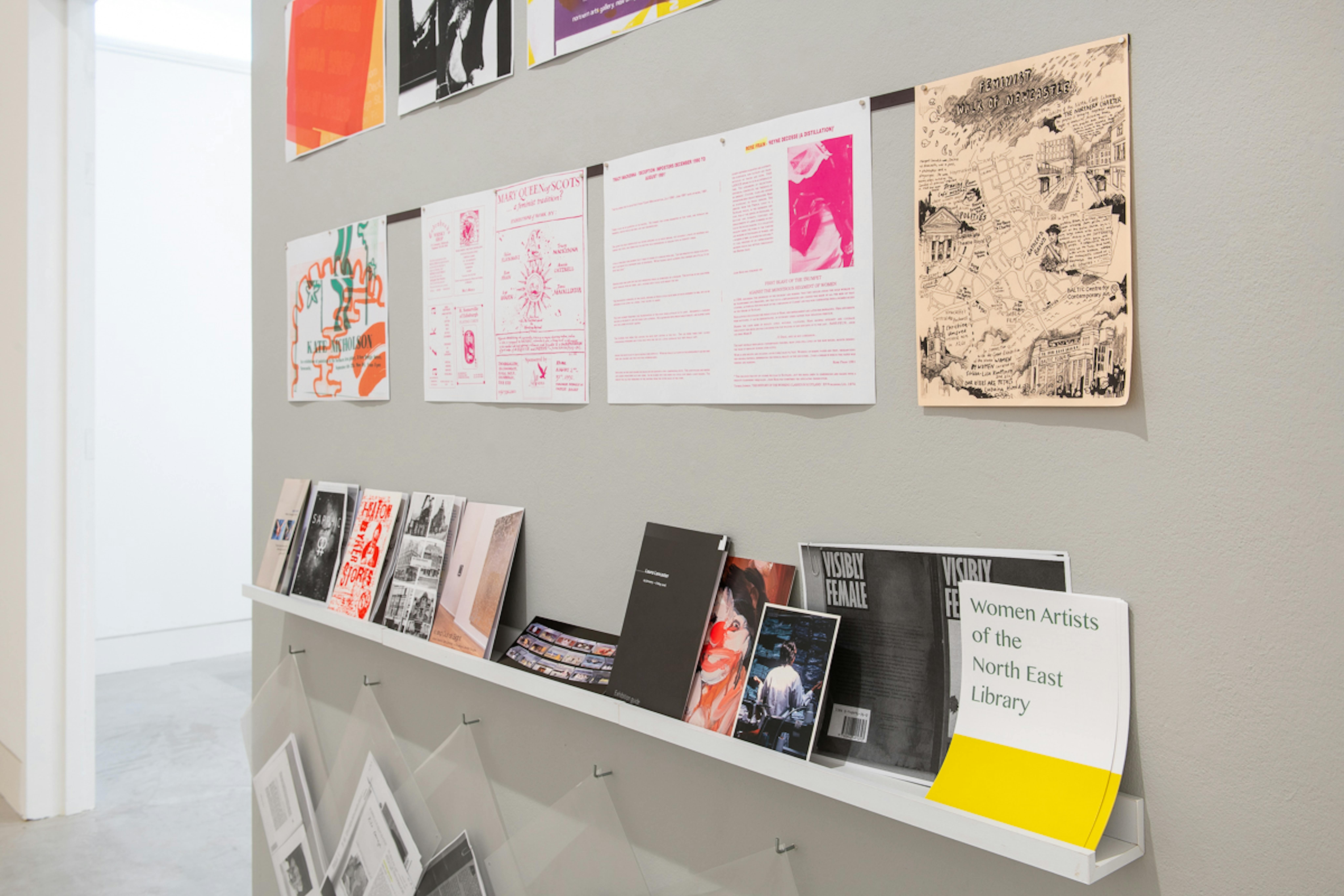 A grey wall contains posters in bright colours taken from an archive of women artists from the North East of England. The shelf contains publications and pamphlets from the archive. 