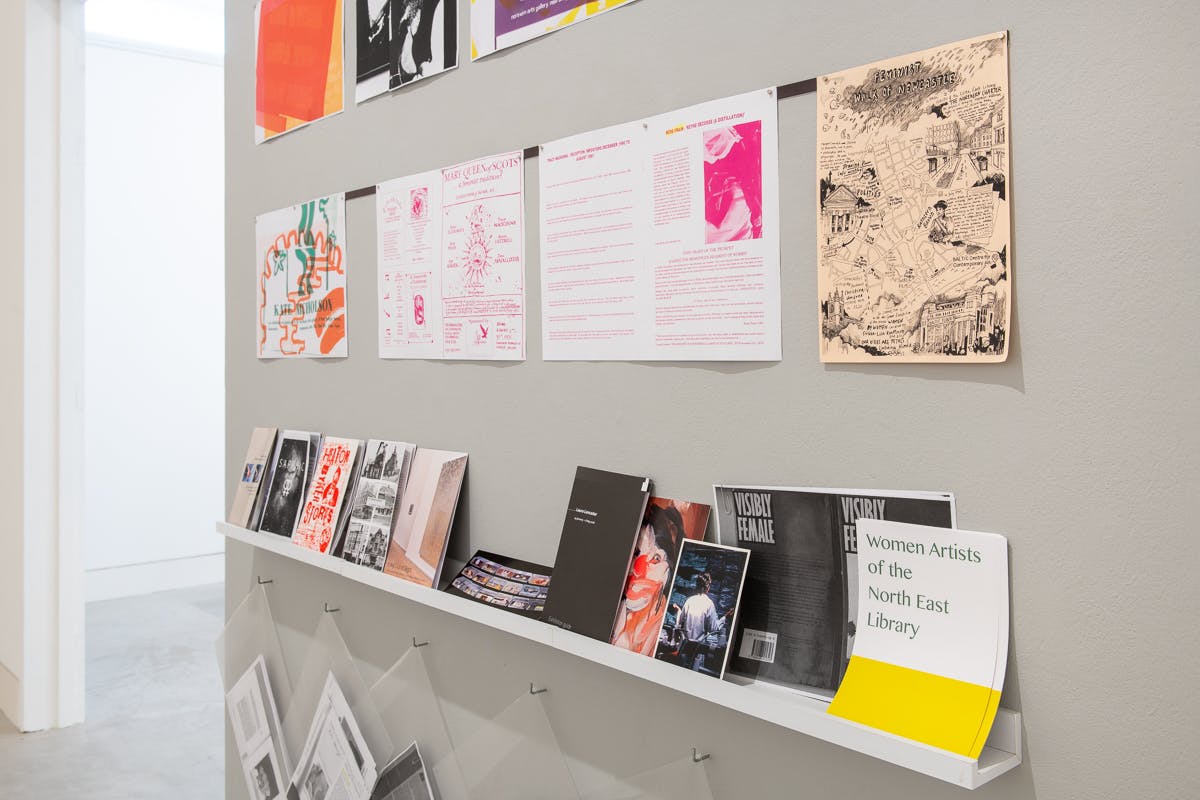 A grey wall contains posters in bright colours taken from an archive of women artists from the North East of England. The shelf contains publications and pamphlets from the archive. 