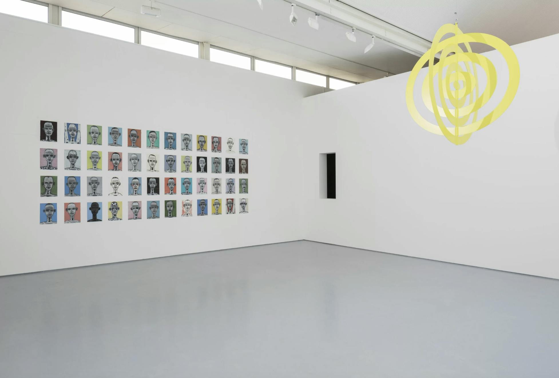 A large white room with light grey floor. On the left hand wall is a series of small simplified portrait paintings arranged in a grid.Hanging from the ceiling is a bright yellow abstract maquette that invokes the sun.
