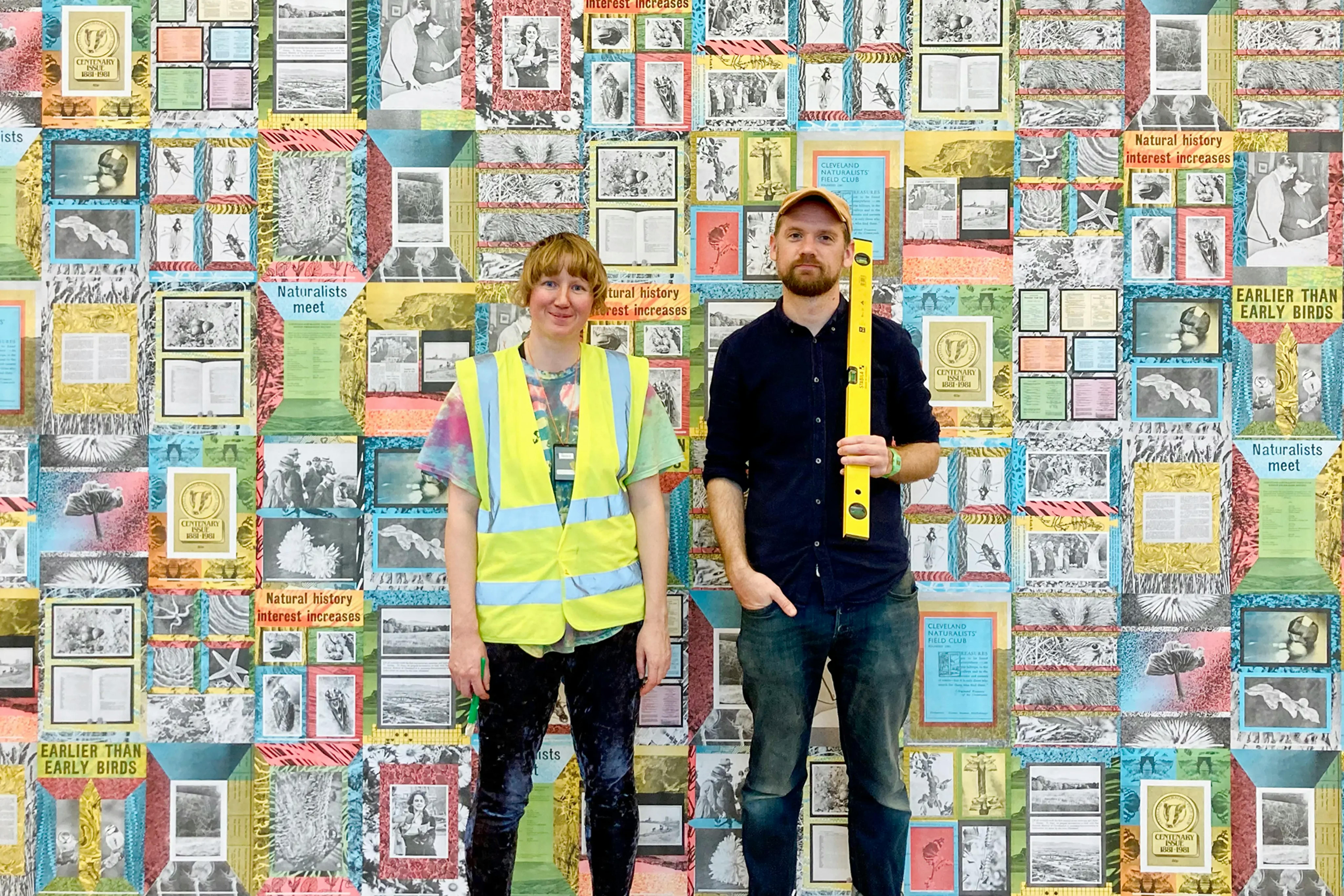 A white woman and man stand in front of a wall of multicoloured posters. The women is wearing a hi-vis jacket and the man, a black shirt, a yellow cap and he is holding a spirit level