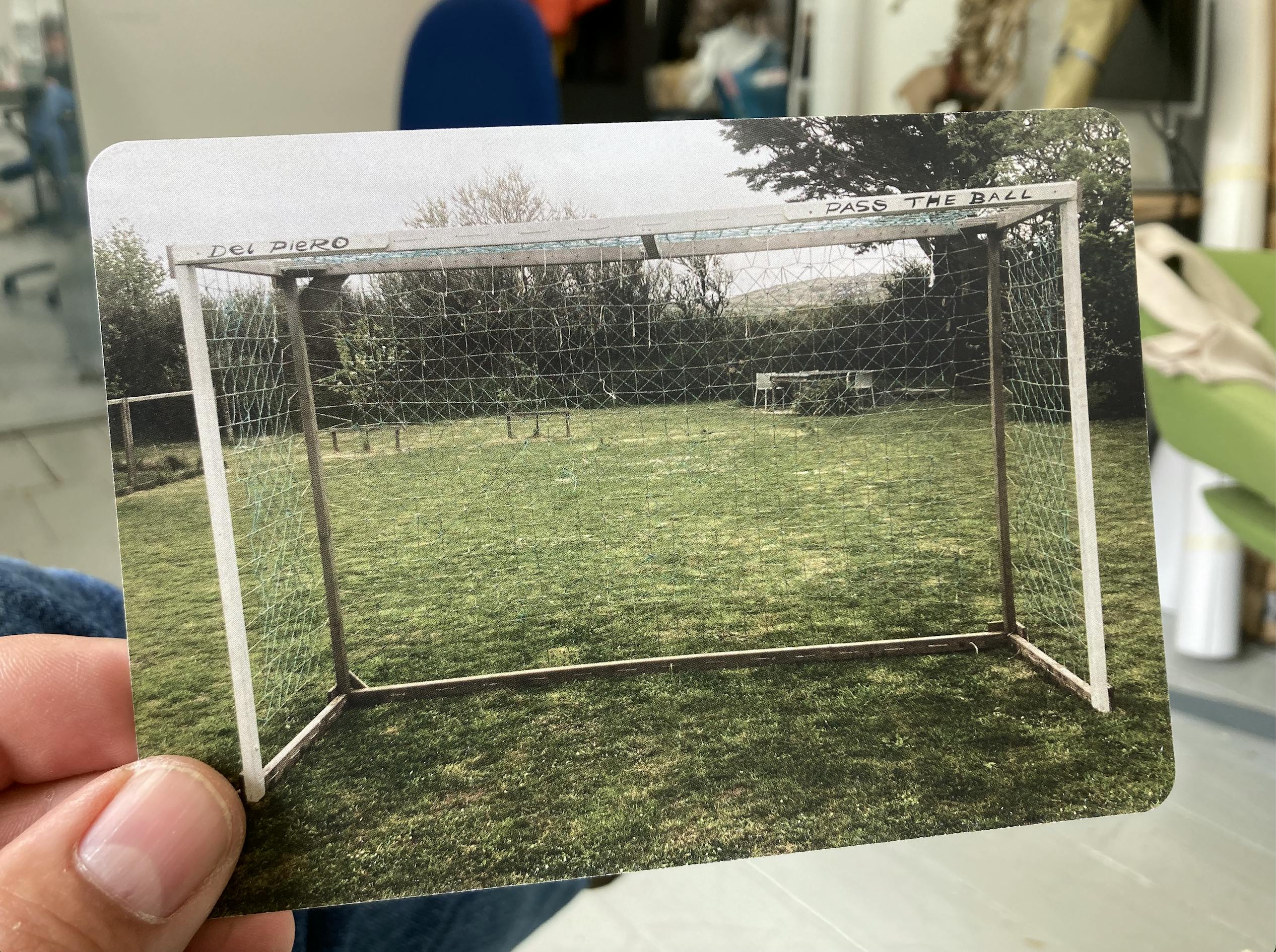 A postcard of a goal frame with a thin white frame. On the top of the frame there is a hand painted sign that says Del Piero Pass the Ball.
