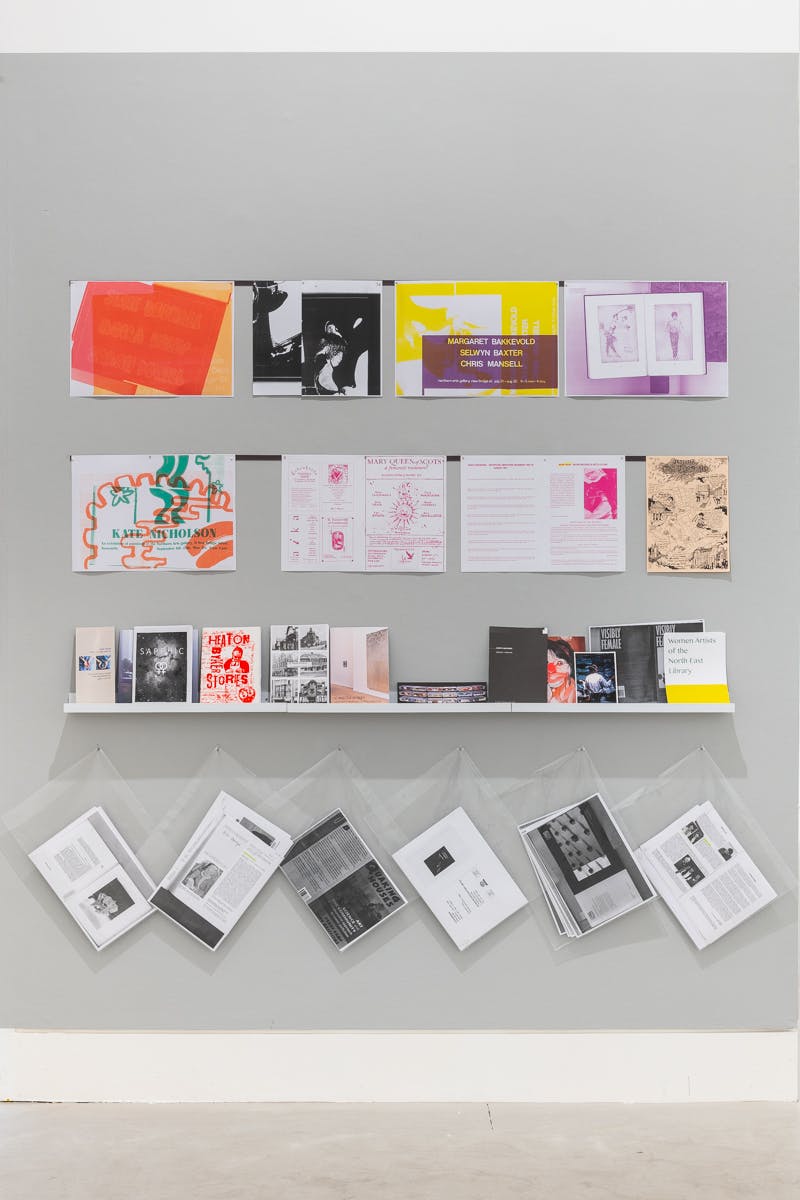 A grey wall contains posters in bright colours taken from an archive of women artists from the North East of England. The shelf contains publications and pamphlets from the archive.
