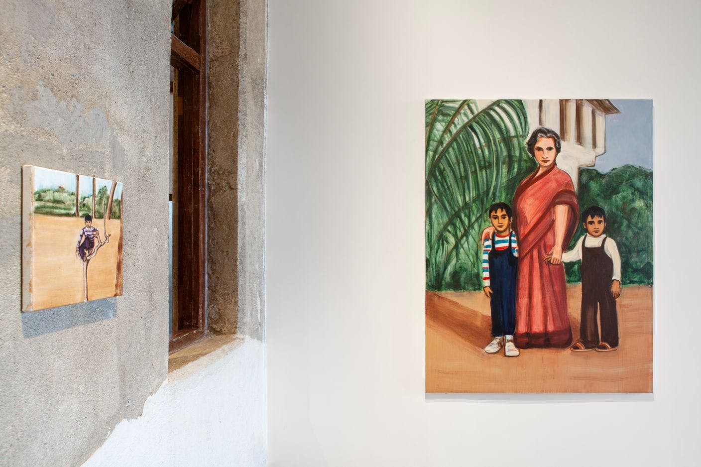 Exhibition view of two figurative paintings. The first on the left features a young boy perched on a tree and the painting on the right features two young brown boys posing with an older white woman. 