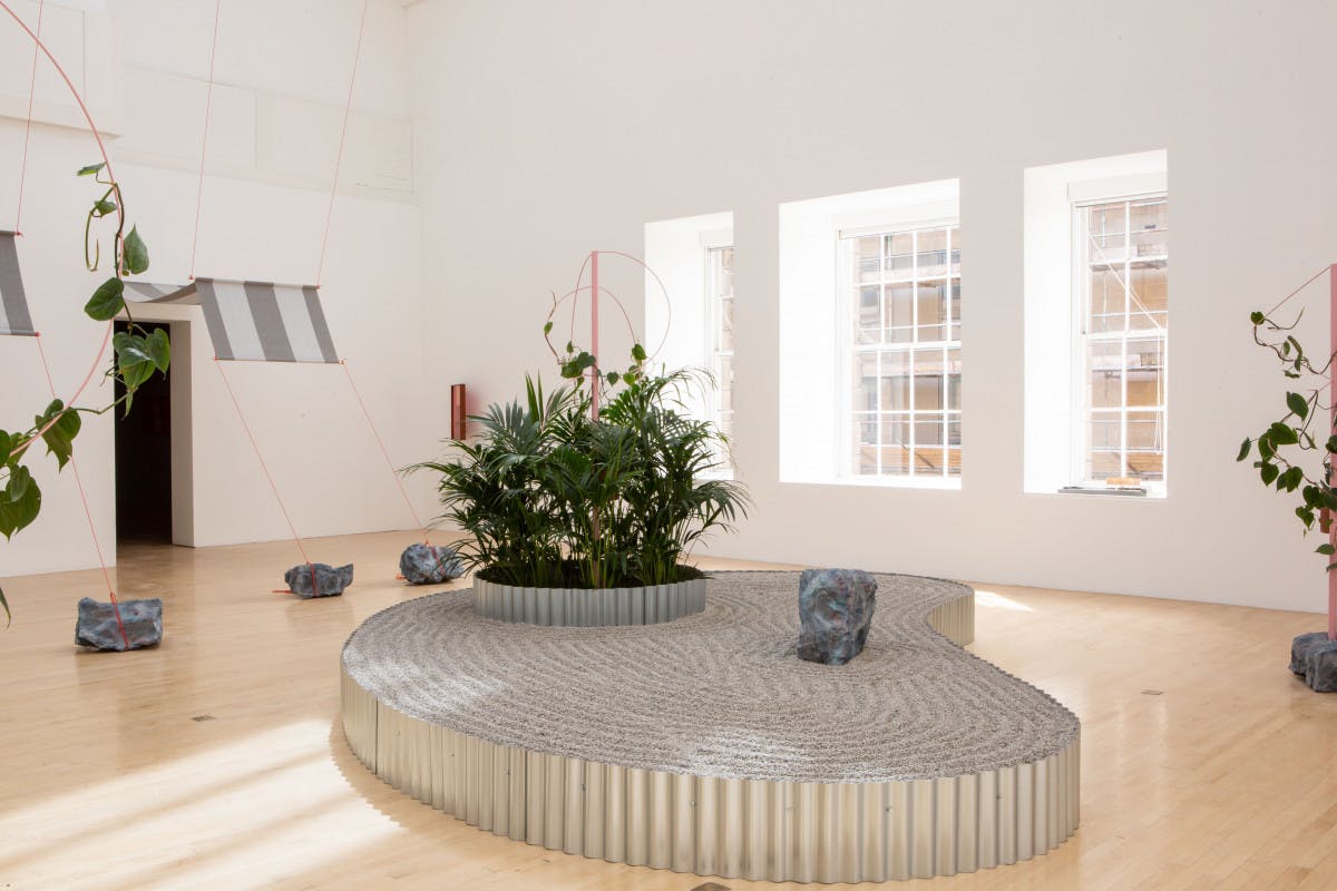 An installation in a bright white gallery. A platform curves around and houses a series of plants. Sculptures are dotted throughout the gallery.