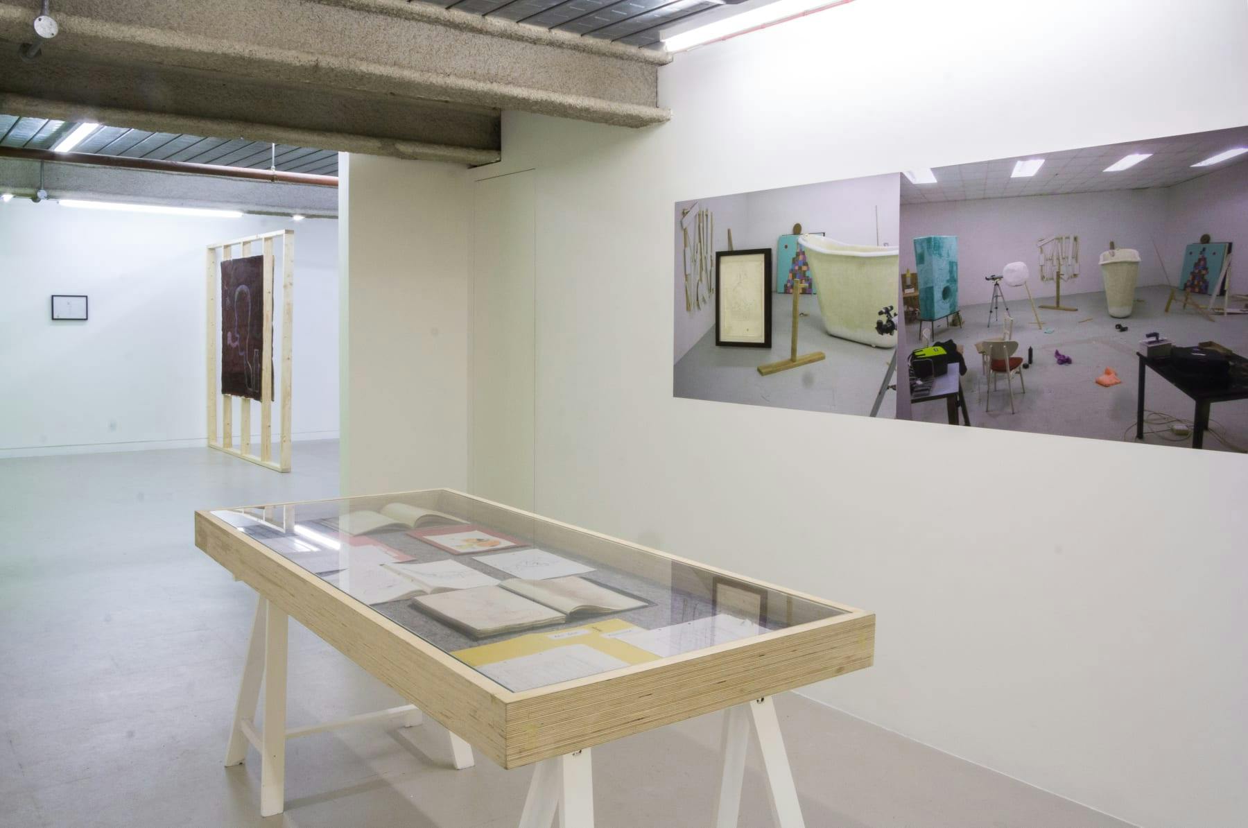 In the foreground is a vitrine with drawings and sketchbooks. The wall contains large photographs of the artist's studio revealing hidden processes that are typically hidden away.